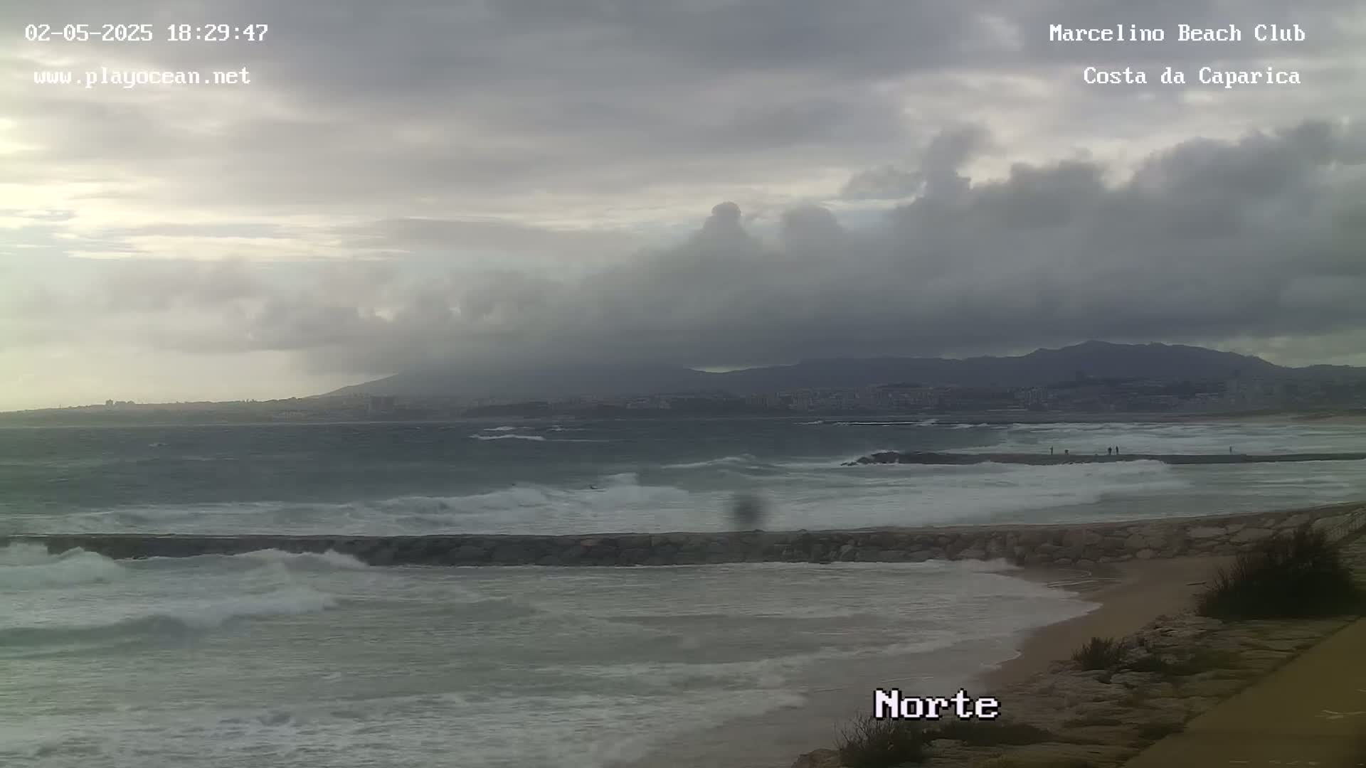 A rocky breakwater protects a sandy beach from choppy waves under a cloudy sky, with distant mountains visible on the horizon.