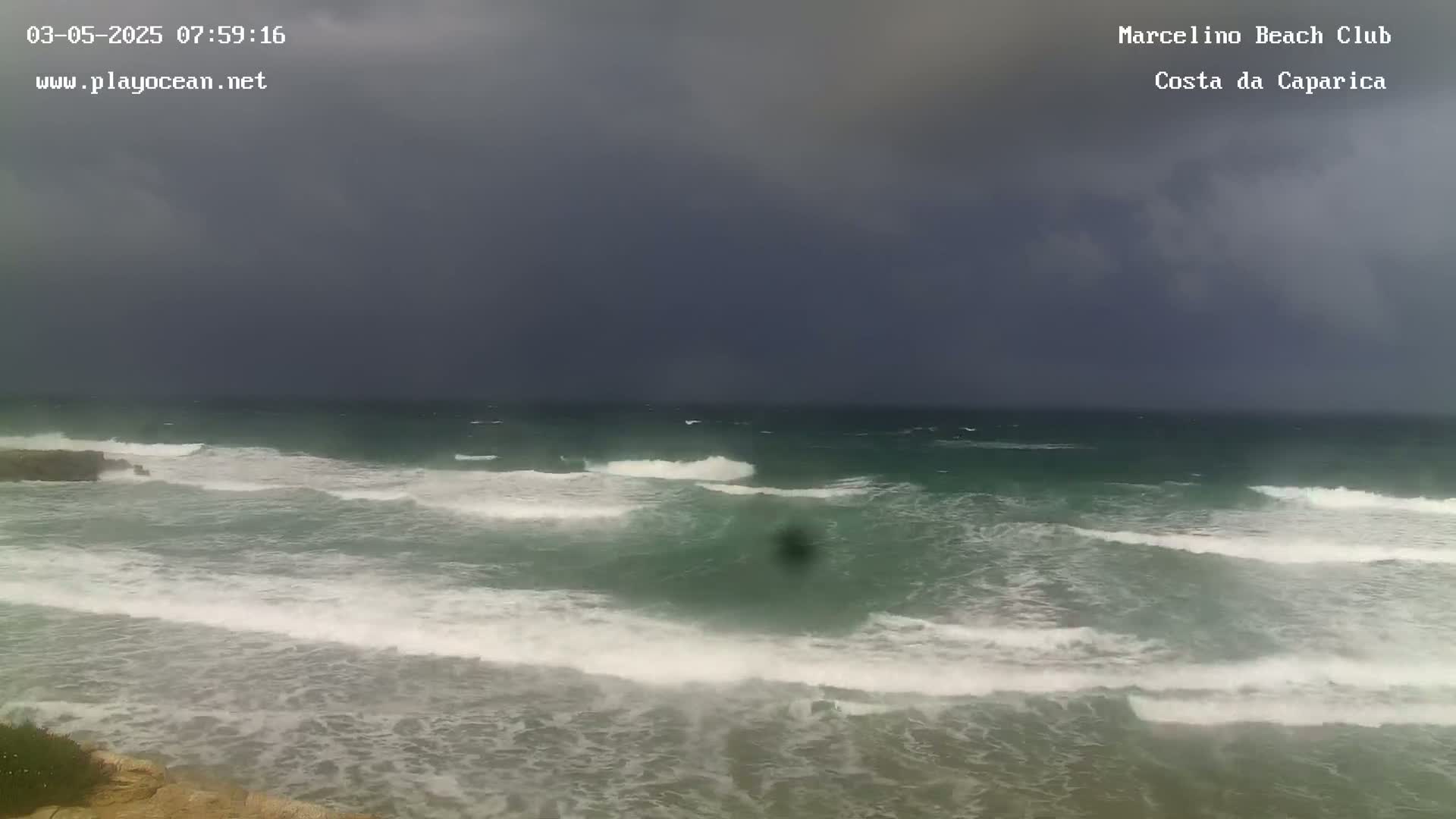 Under a dark, overcast sky, moderately sized waves crash on a sandy beach.