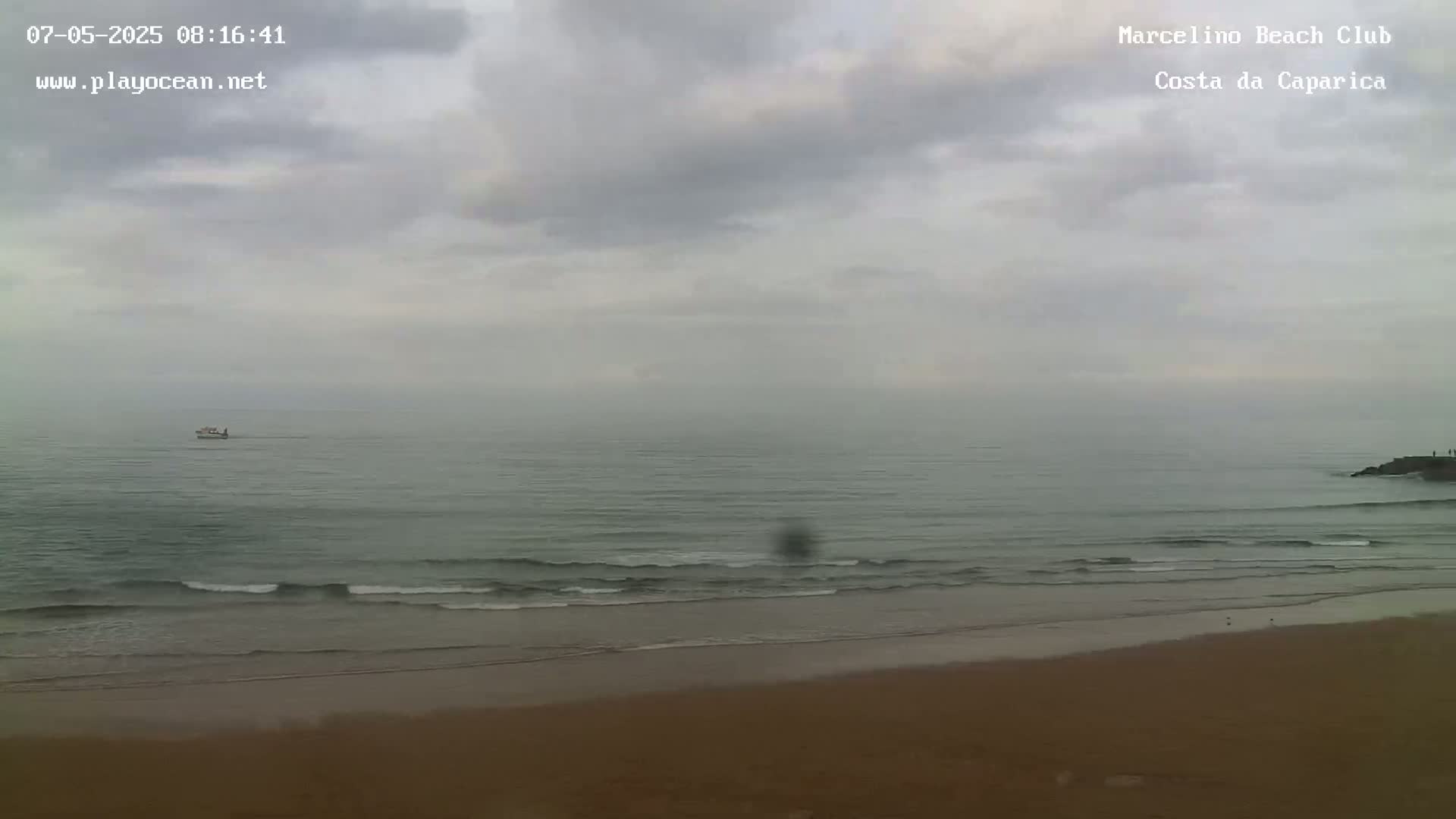 A hazy, overcast day at a beach shows a calm ocean with small waves lapping a sandy shore, and a distant small boat.