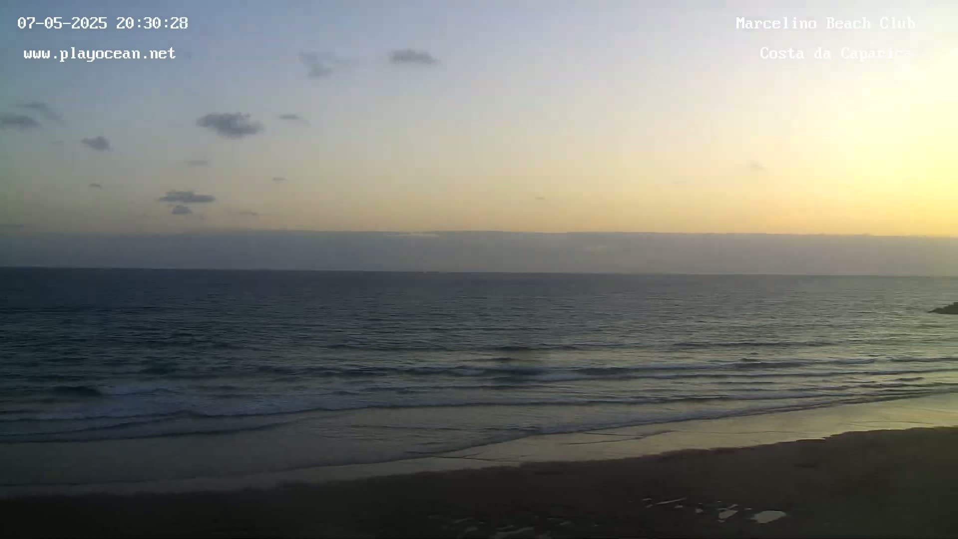 A mostly clear, sunset sky over a dark ocean with gentle waves lapping a sandy beach.