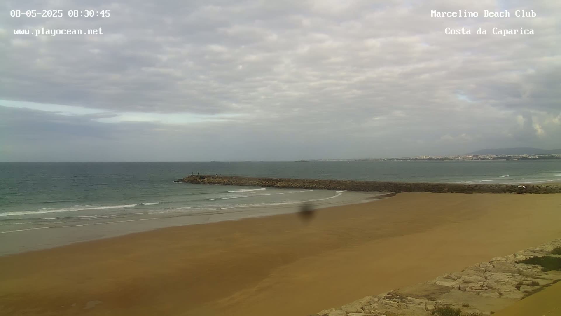 A sandy beach with a rock jetty extends into a calm, overcast ocean, with a distant shoreline visible in the background.