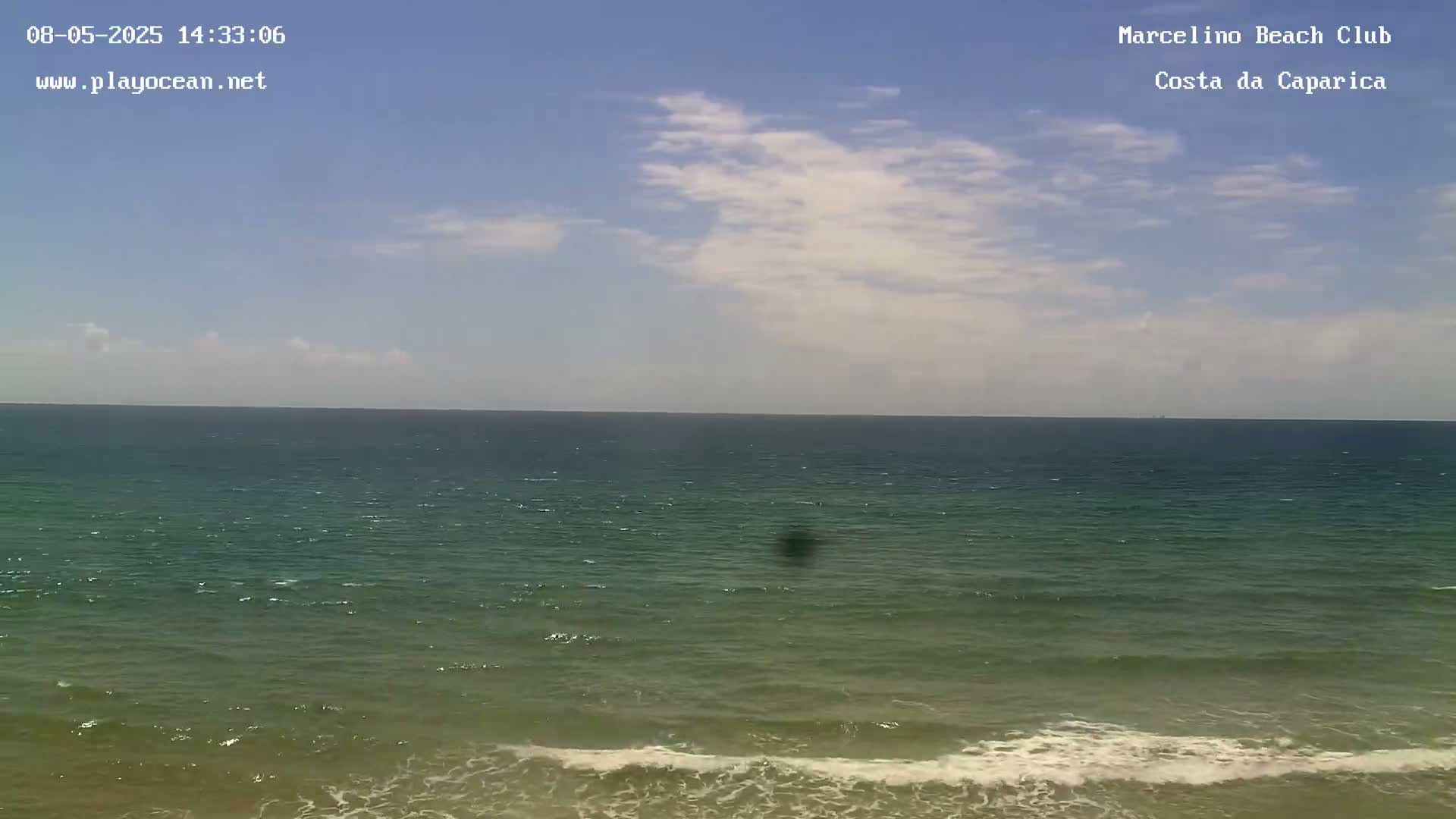 A mostly sunny day shows a wide expanse of ocean with small waves lapping the shore under a partly cloudy sky.
