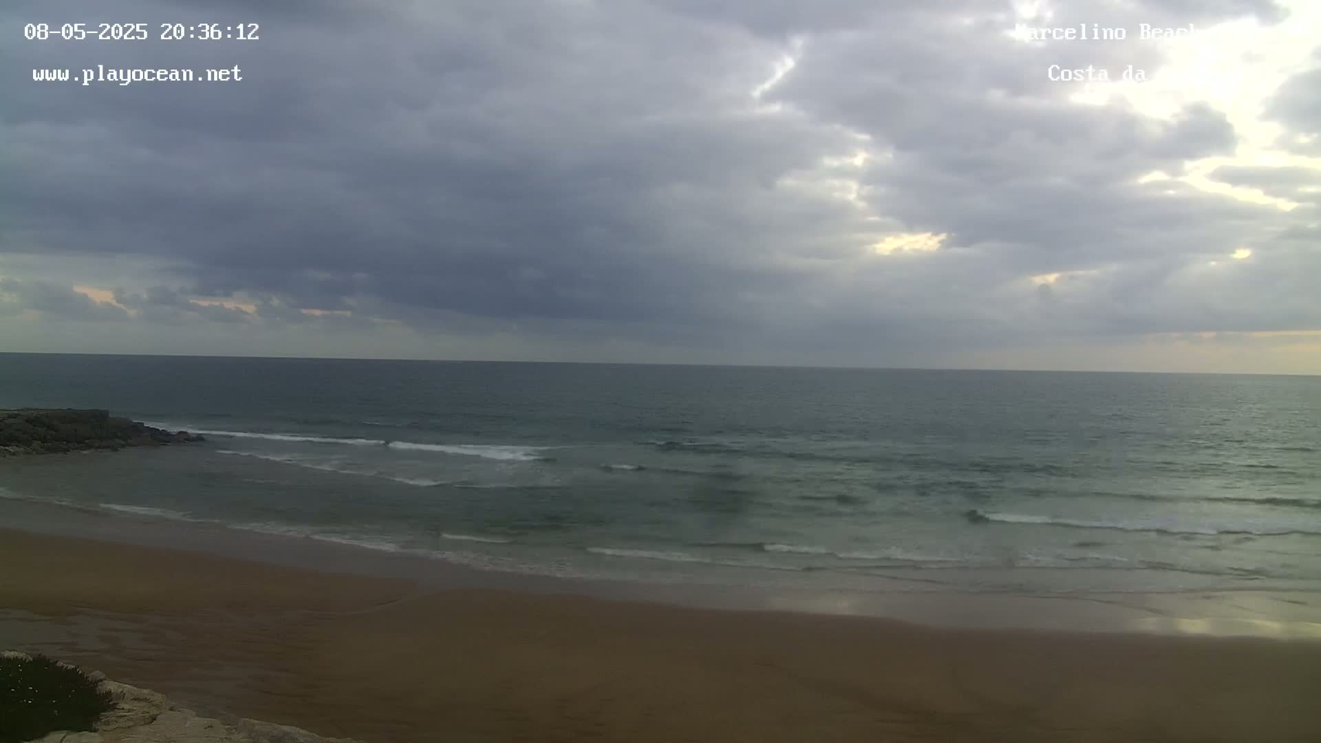 A sandy beach with gentle waves rolls onto the shore under a cloudy, overcast sky.