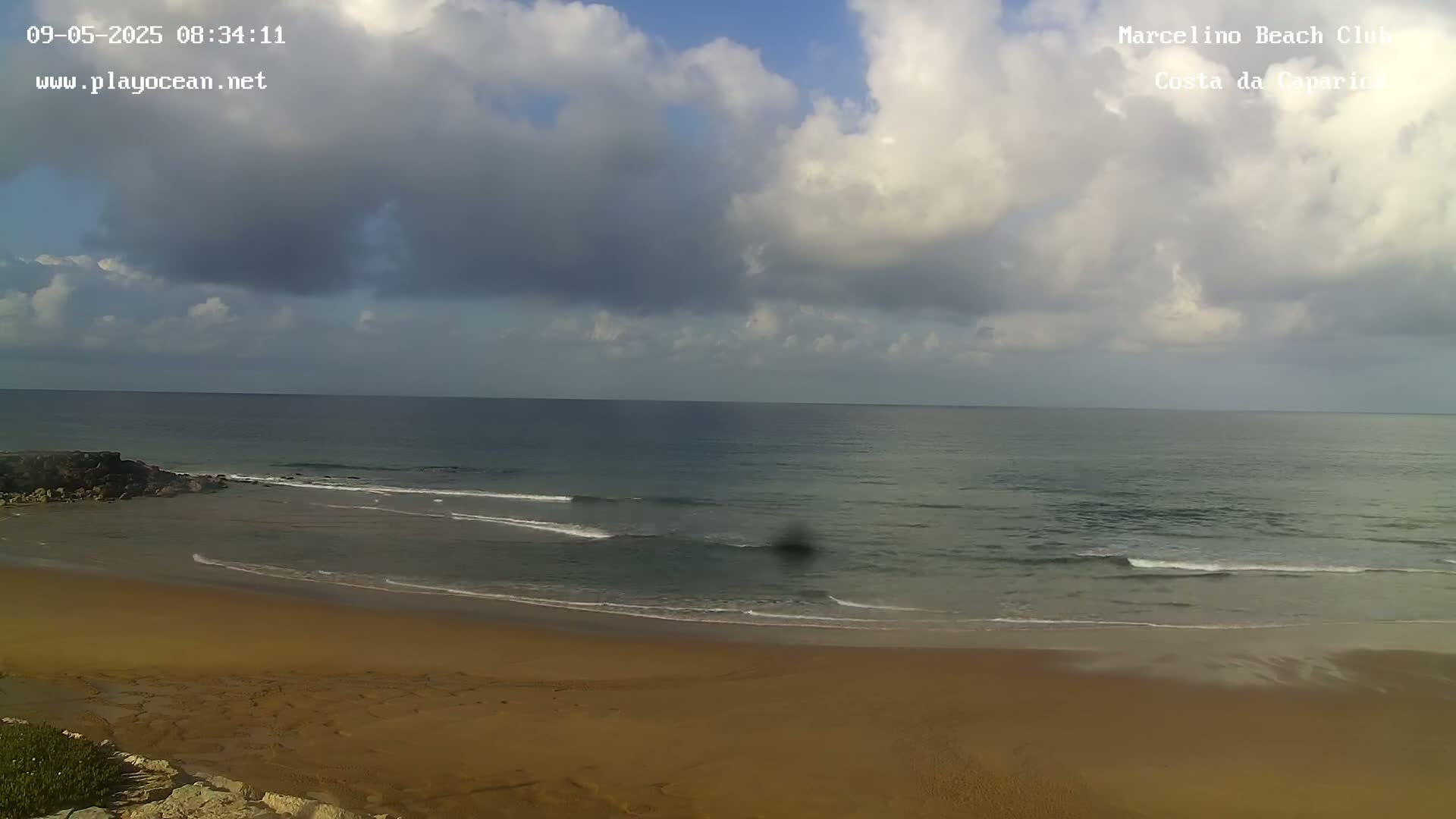 A partly cloudy sky overlooks a sandy beach and calm ocean with small waves.