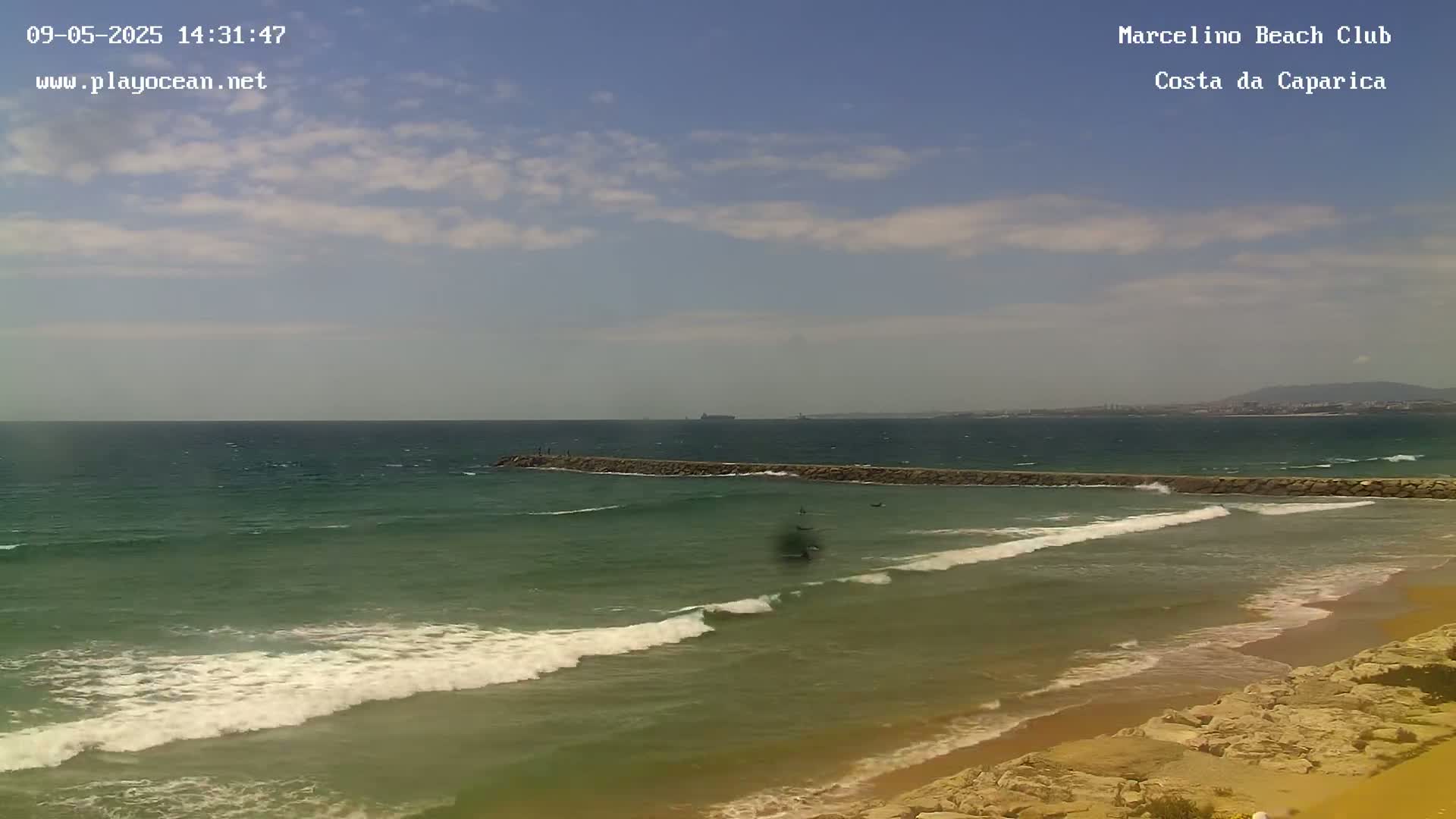 A partly cloudy day shows moderate waves breaking on a sandy beach with a long rock jetty extending into the ocean.