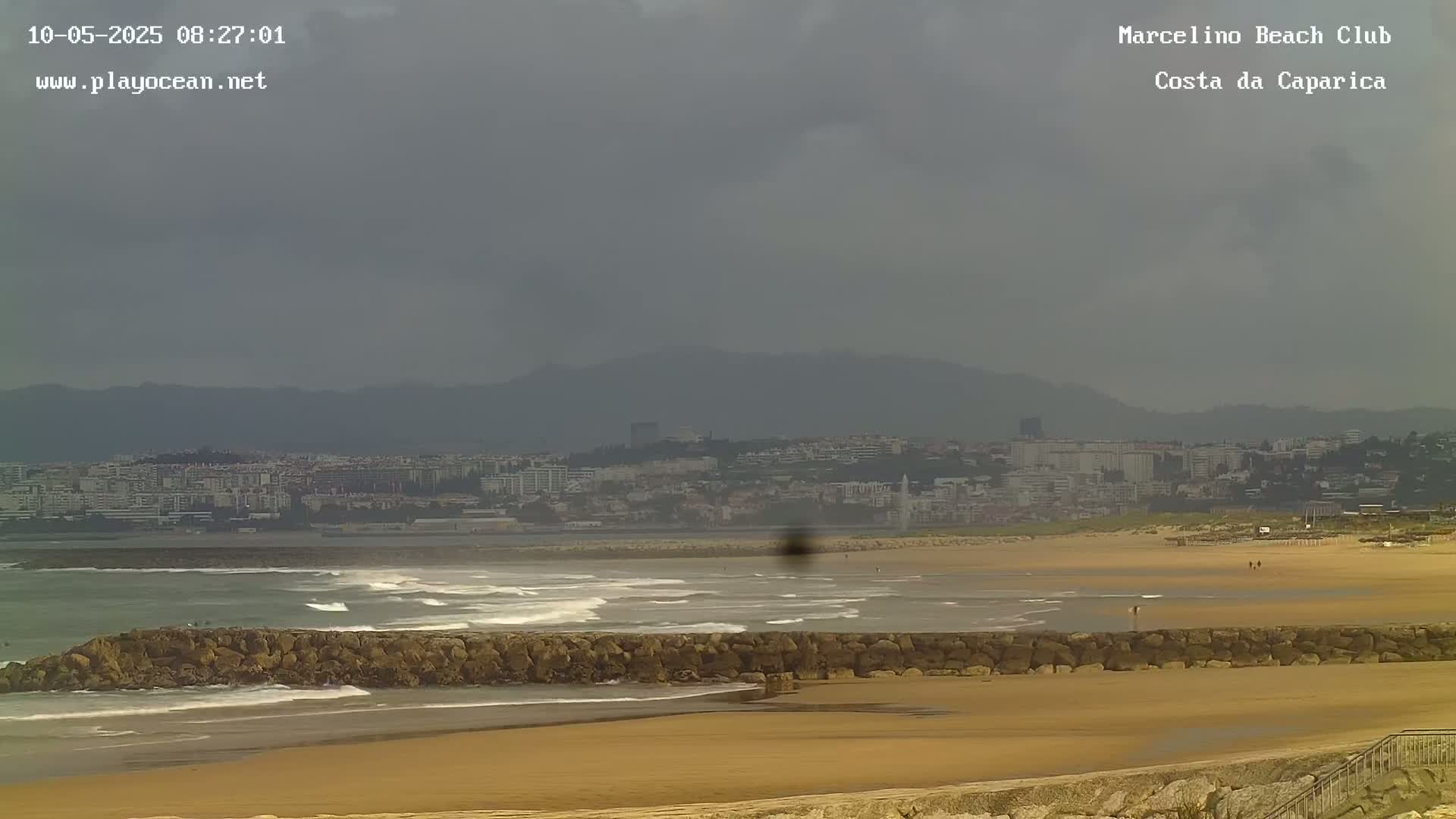 A sandy beach with small waves and a rock wall stretches out before a distant city under an overcast sky.