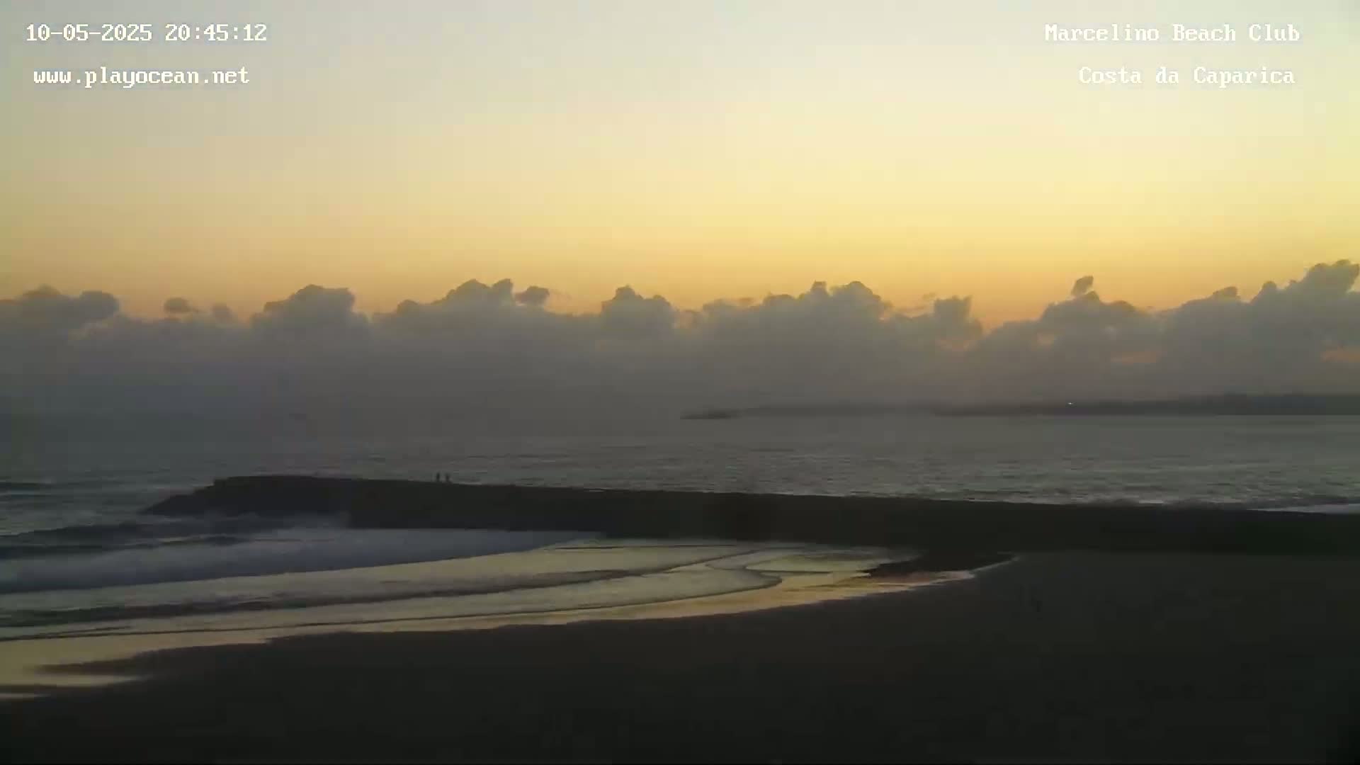 A beach at sunset with calm waves breaking gently on the shore under a mostly cloudy, pale orange sky.