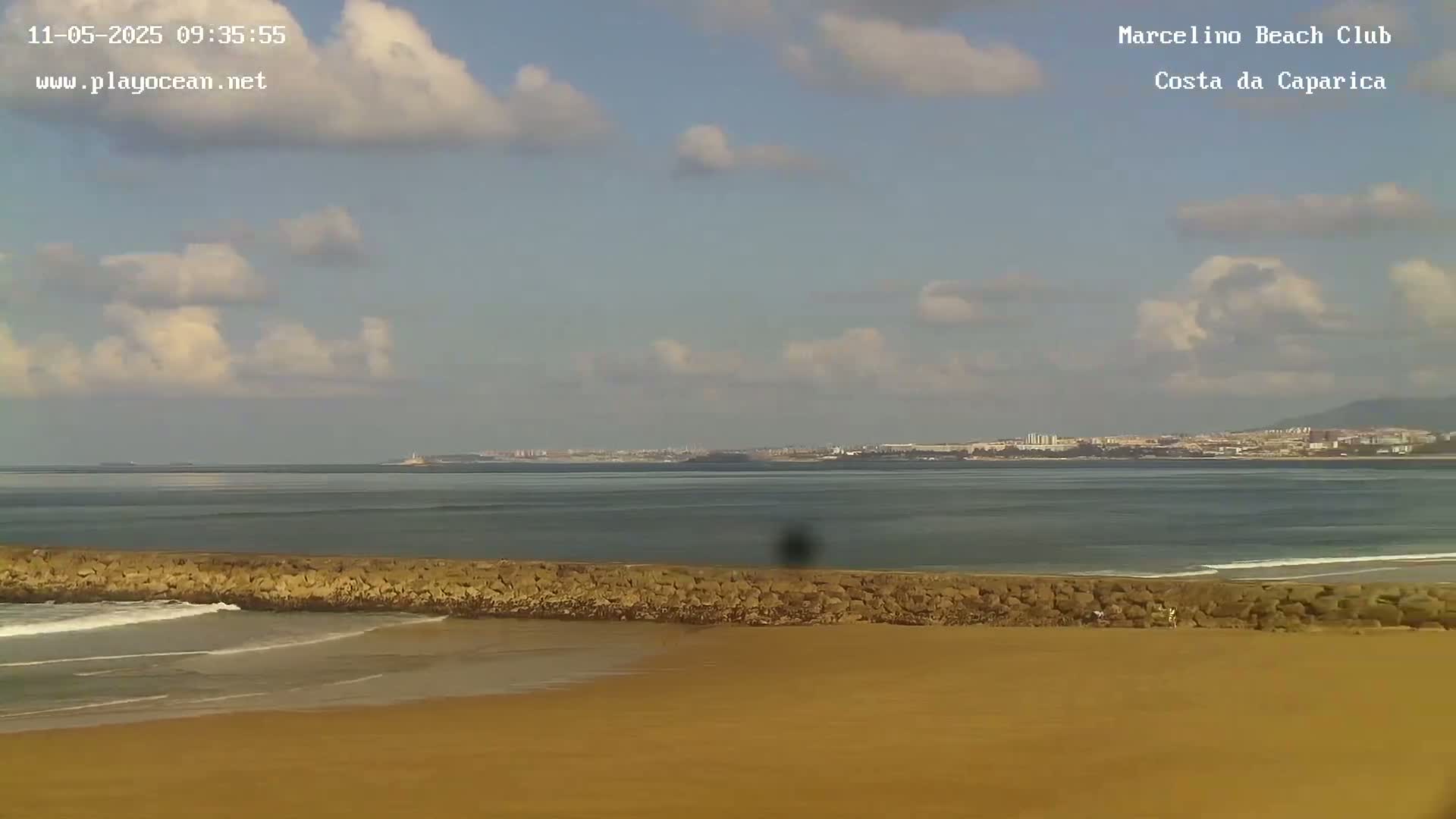 A sandy beach with a low rock wall separating it from a calm ocean under a partly cloudy sky.