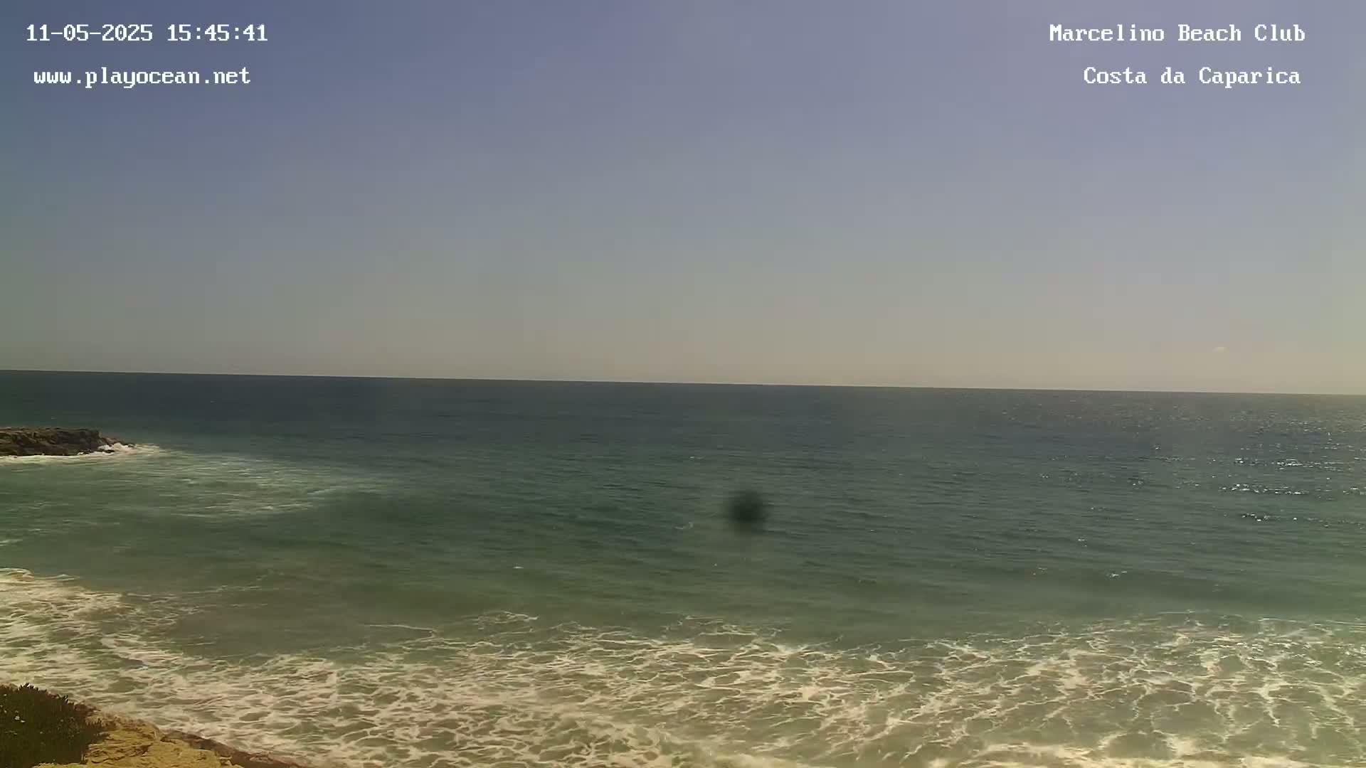 A mostly sunny day shows a calm ocean with small waves breaking on a sandy shore.