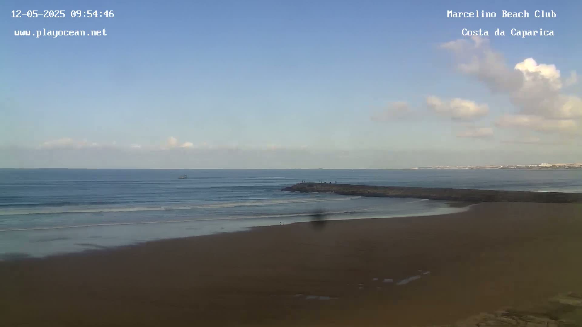 A sandy beach with small waves under a partly cloudy sky meets a dark rock jetty extending into a calm ocean.