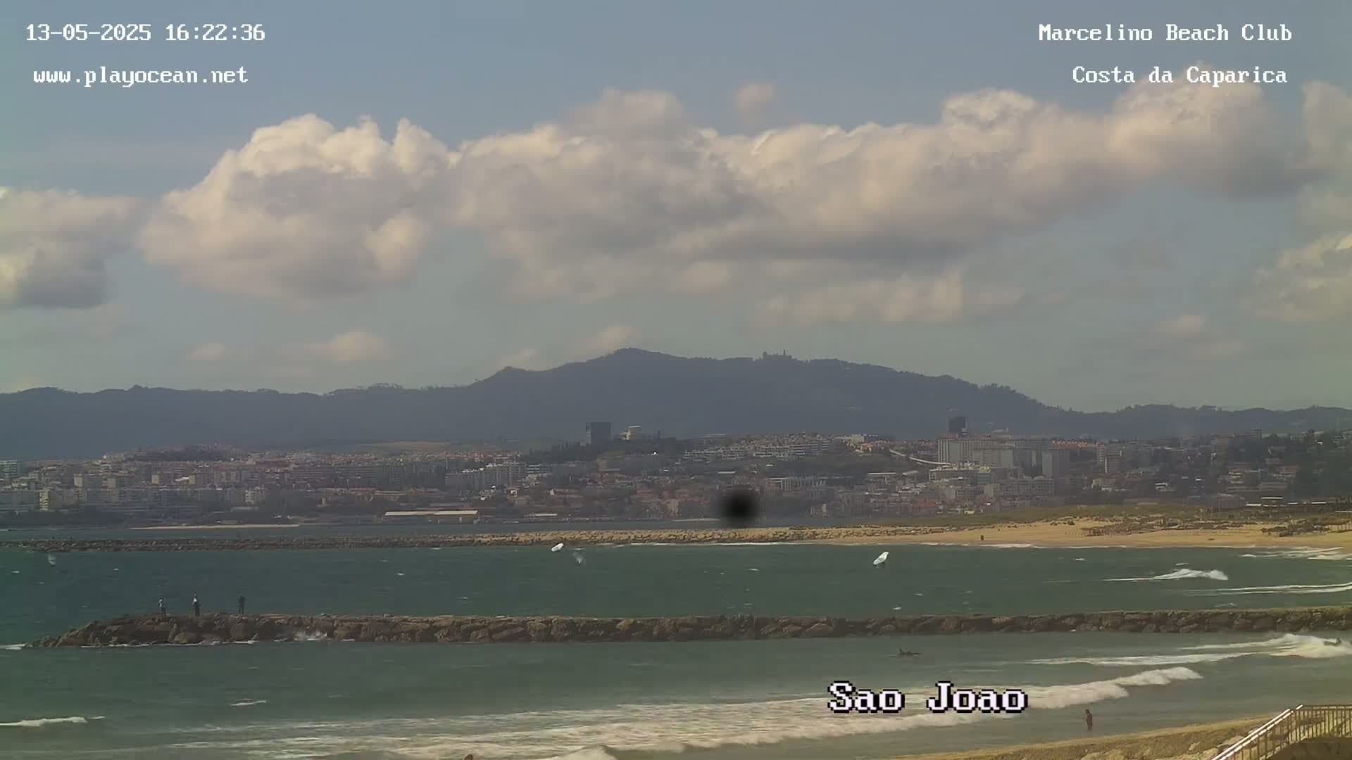 A partly cloudy day shows a beach with a rocky breakwater, a calm ocean with a few small waves, and a distant cityscape nestled against mountains.