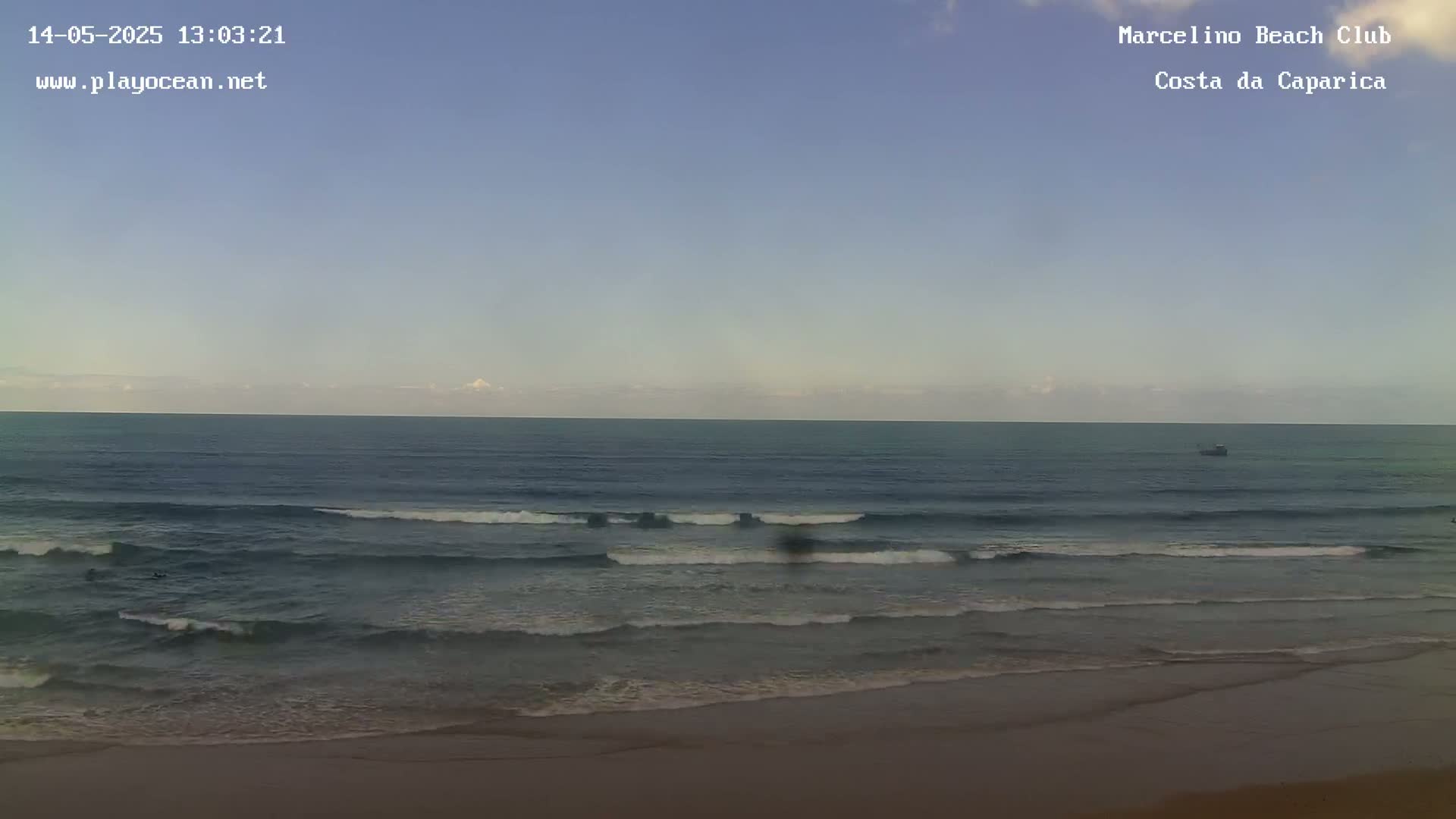 A sandy beach meets a calm ocean under a clear, light-blue sky with a few small clouds on the horizon.