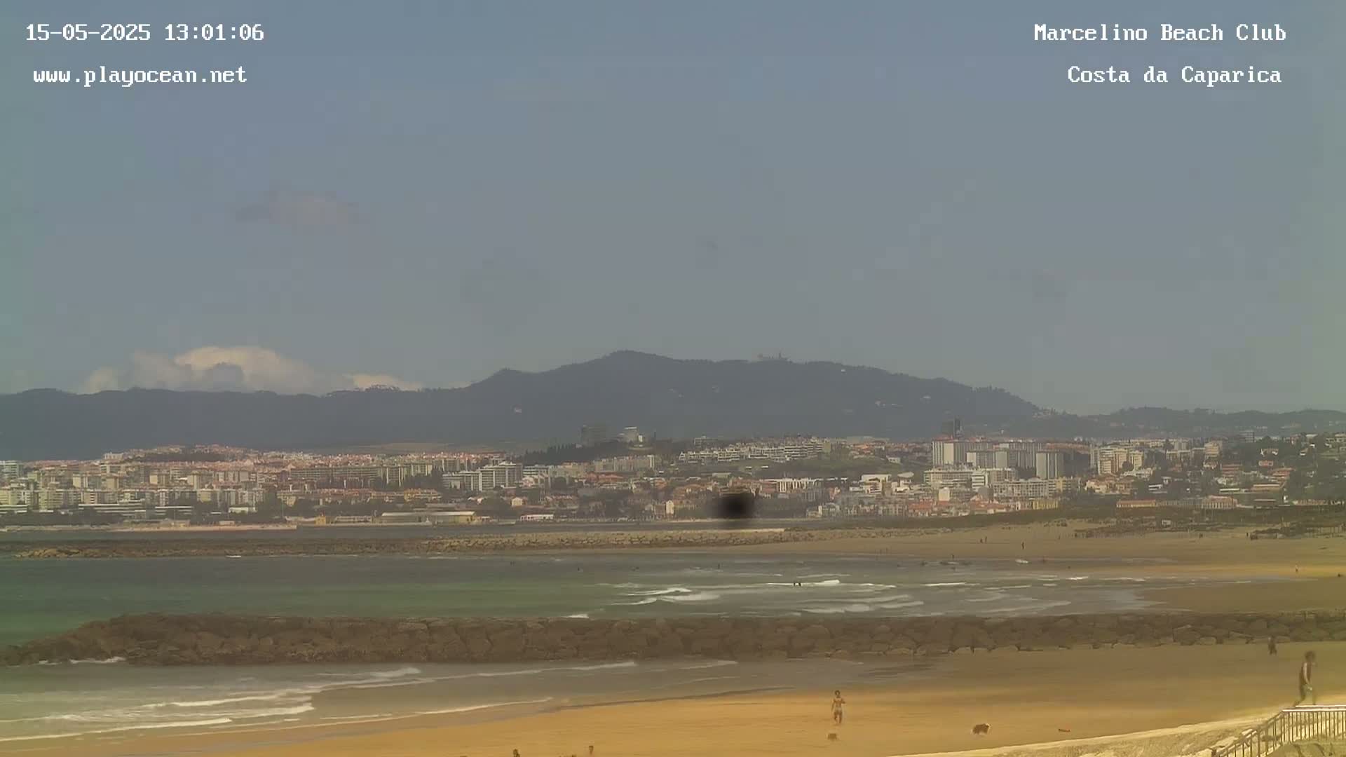 A sandy beach with gentle waves laps the shore, a rocky breakwater sits in the foreground, and a distant town is visible beneath hazy mountains under a mostly clear sky.
