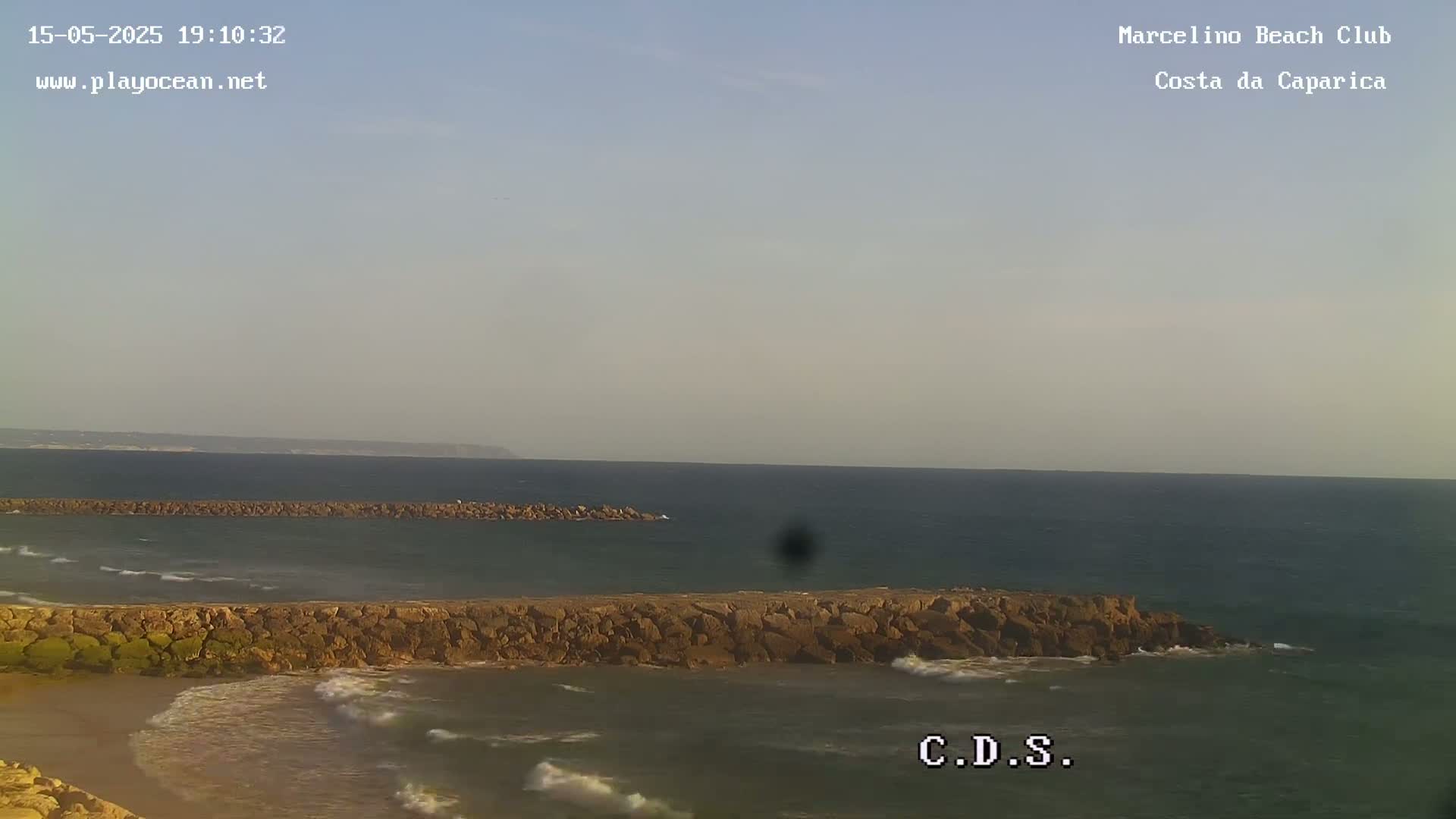 A rocky breakwater separates a calm, somewhat overcast ocean from a sandy beach.