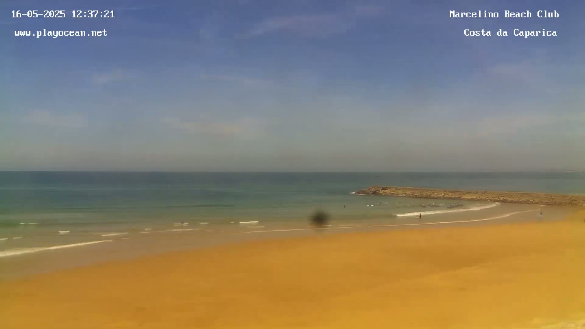 A sandy beach slopes down to a calm ocean with a few surfers visible near a rock jetty under a clear, sunny sky.