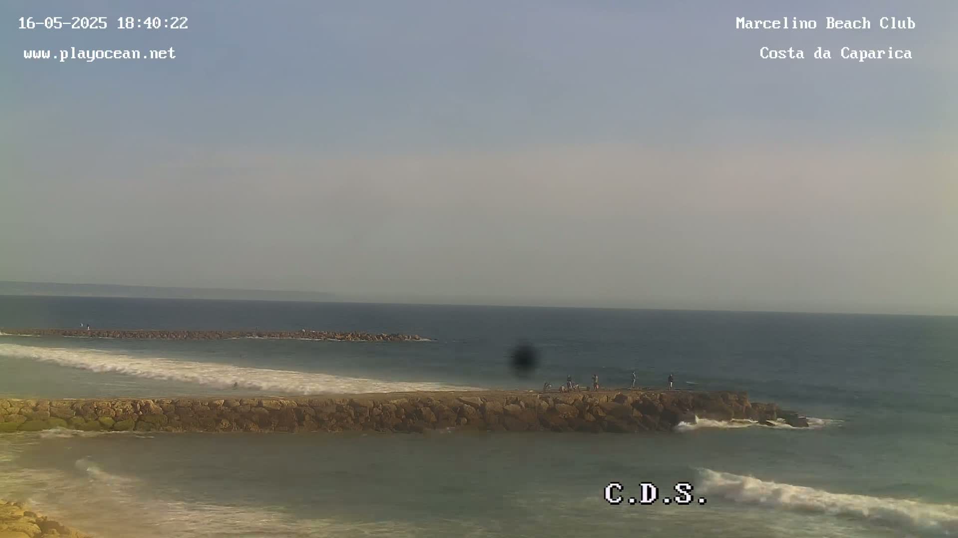 A rocky jetty extends into a calm ocean under a hazy sky, with a few people visible on the rocks.