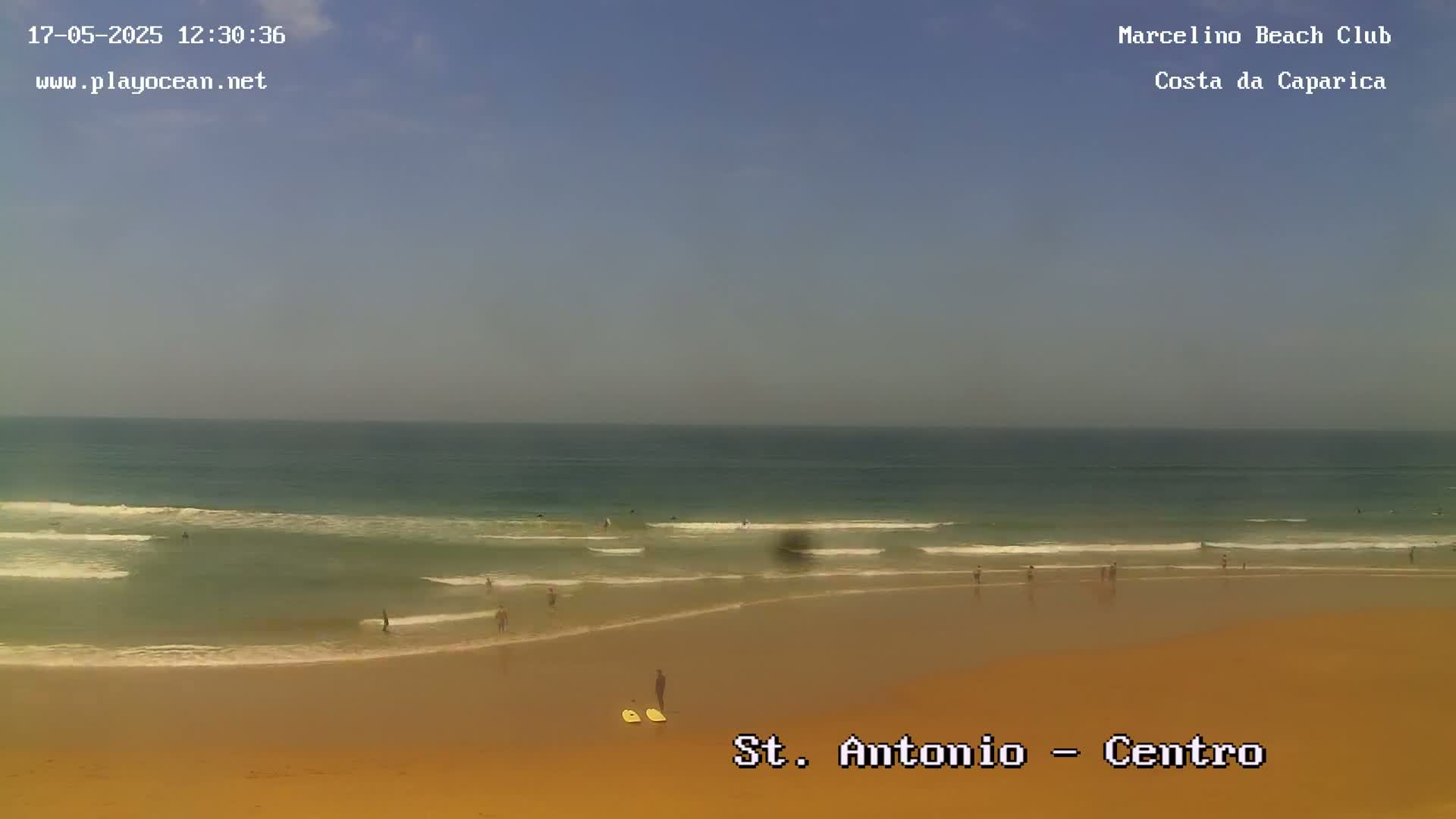 A sandy beach with a few people on it and small waves rolling onto the shore under a hazy sky.