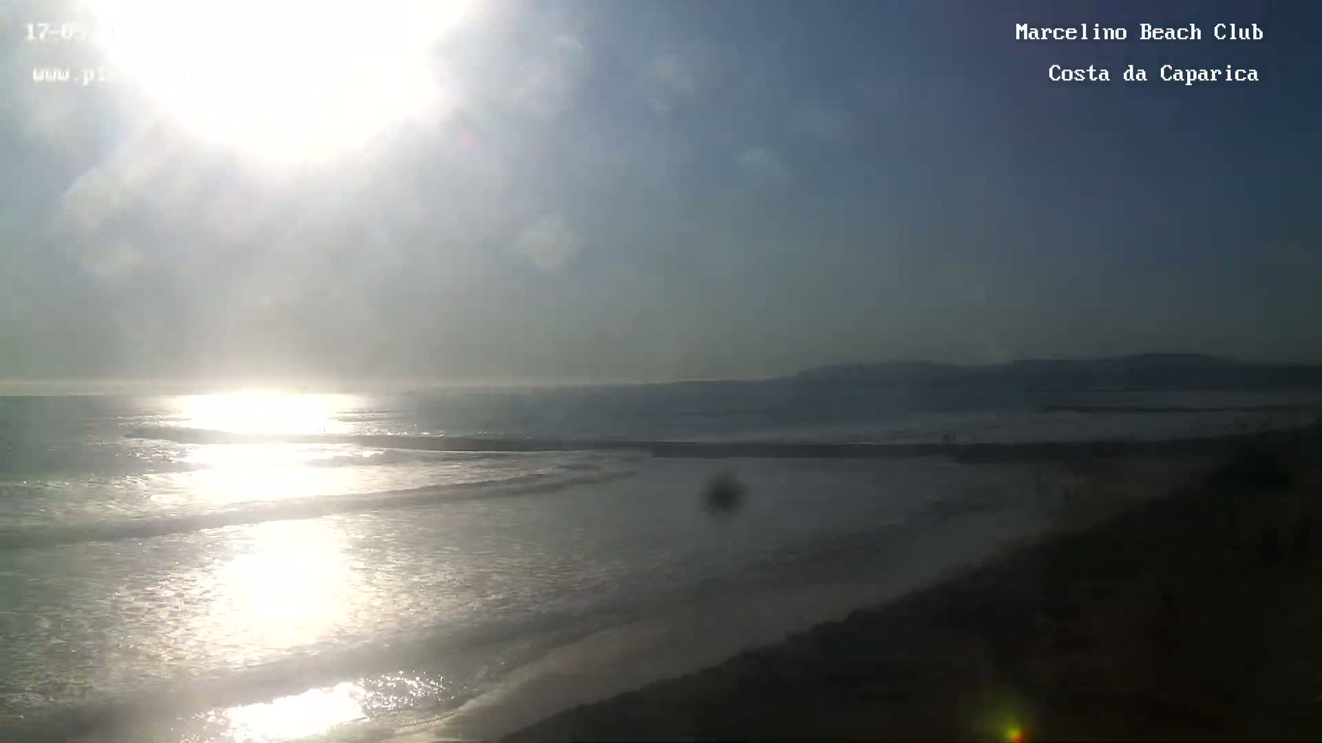 A bright, sunny day shows a wide view of an ocean beach with small waves gently lapping the shore.