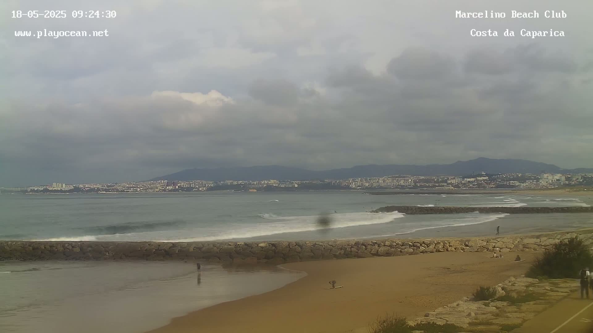 A sandy beach with a stone wall separating it from a wavy ocean, under a cloudy sky, shows a distant city and mountains in the background.