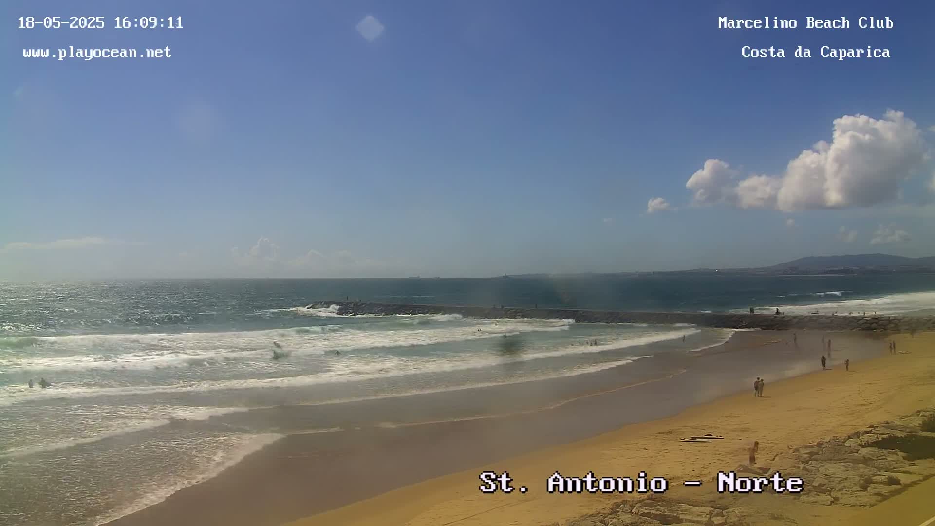 A sandy beach with people on it is shown, with waves breaking on the shore under a partly cloudy sky.