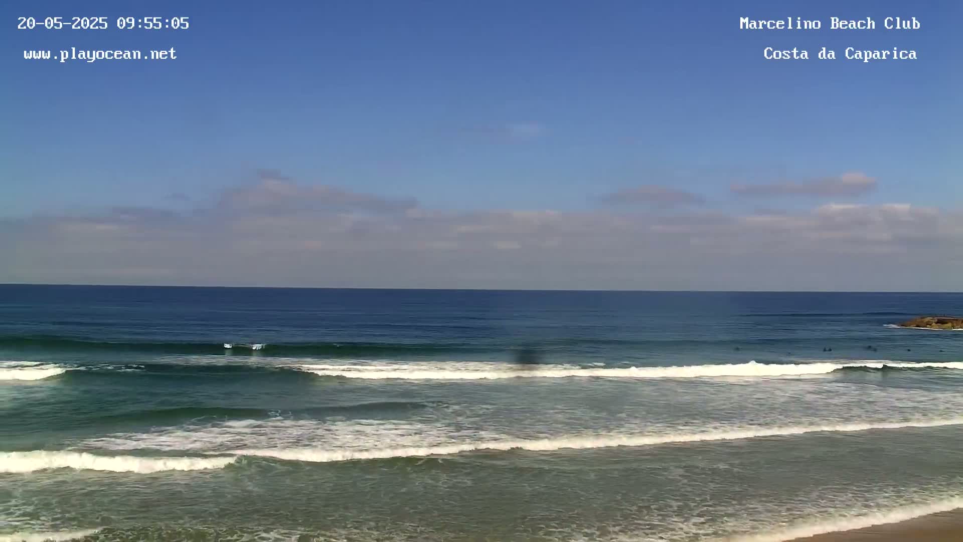 Under a mostly clear blue sky, several surfers ride waves that gently roll onto a sandy beach.