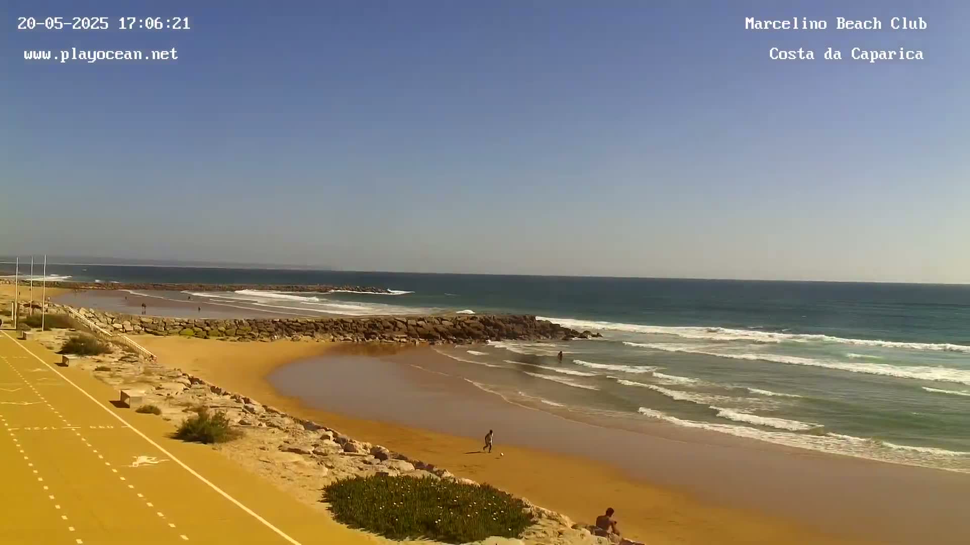 A sandy beach with a few people on it and small waves lapping the shore under a clear blue sky.
