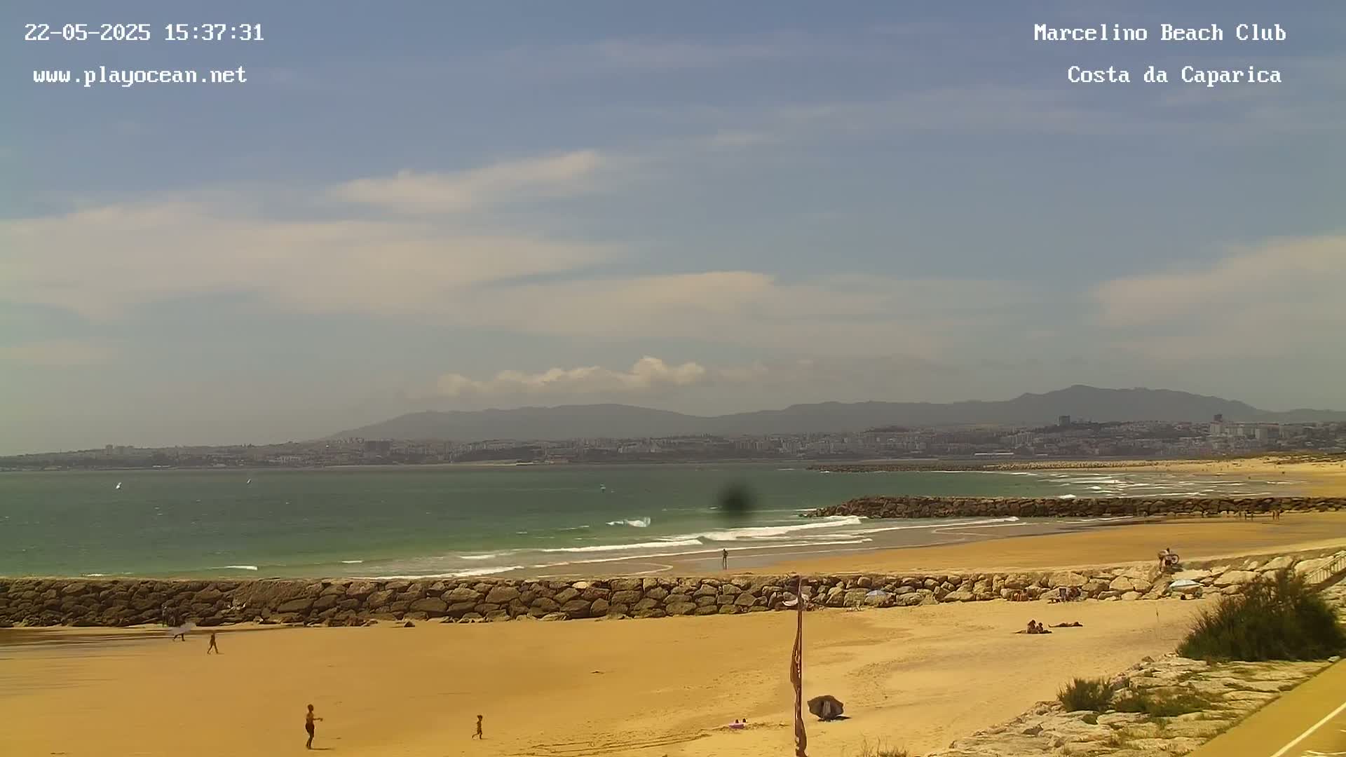 A sandy beach with a rock wall, calm ocean waves, and a few people enjoying a partly sunny day with some clouds.