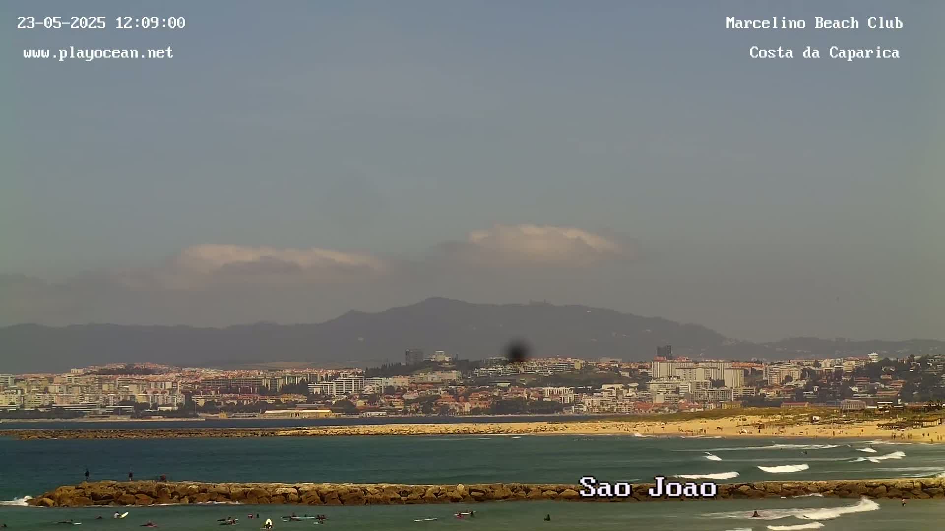 A hazy, partly cloudy day shows a beach with small waves, surfers in the water, and a coastal city in the background, with mountains visible in the distance.