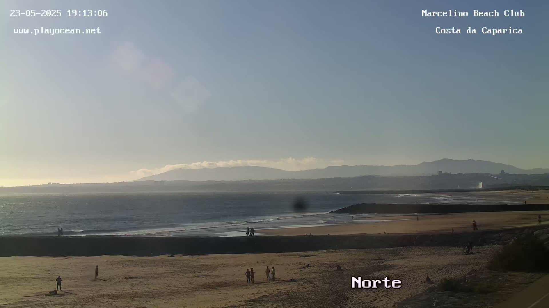 A sandy beach with a few people on it stretches to a calm ocean under a clear, light-blue sky with distant mountains visible on the horizon.