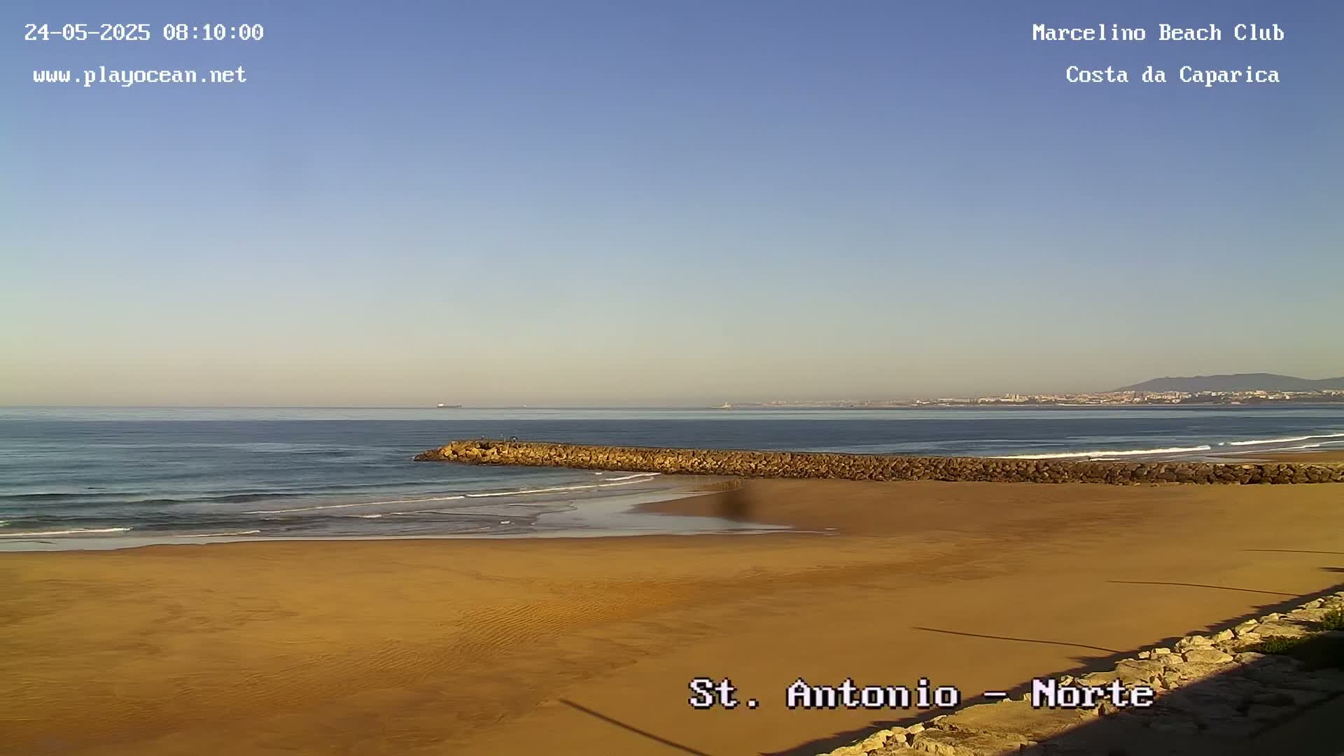 A sandy beach with a rock jetty extends into a calm ocean under a clear blue sky.