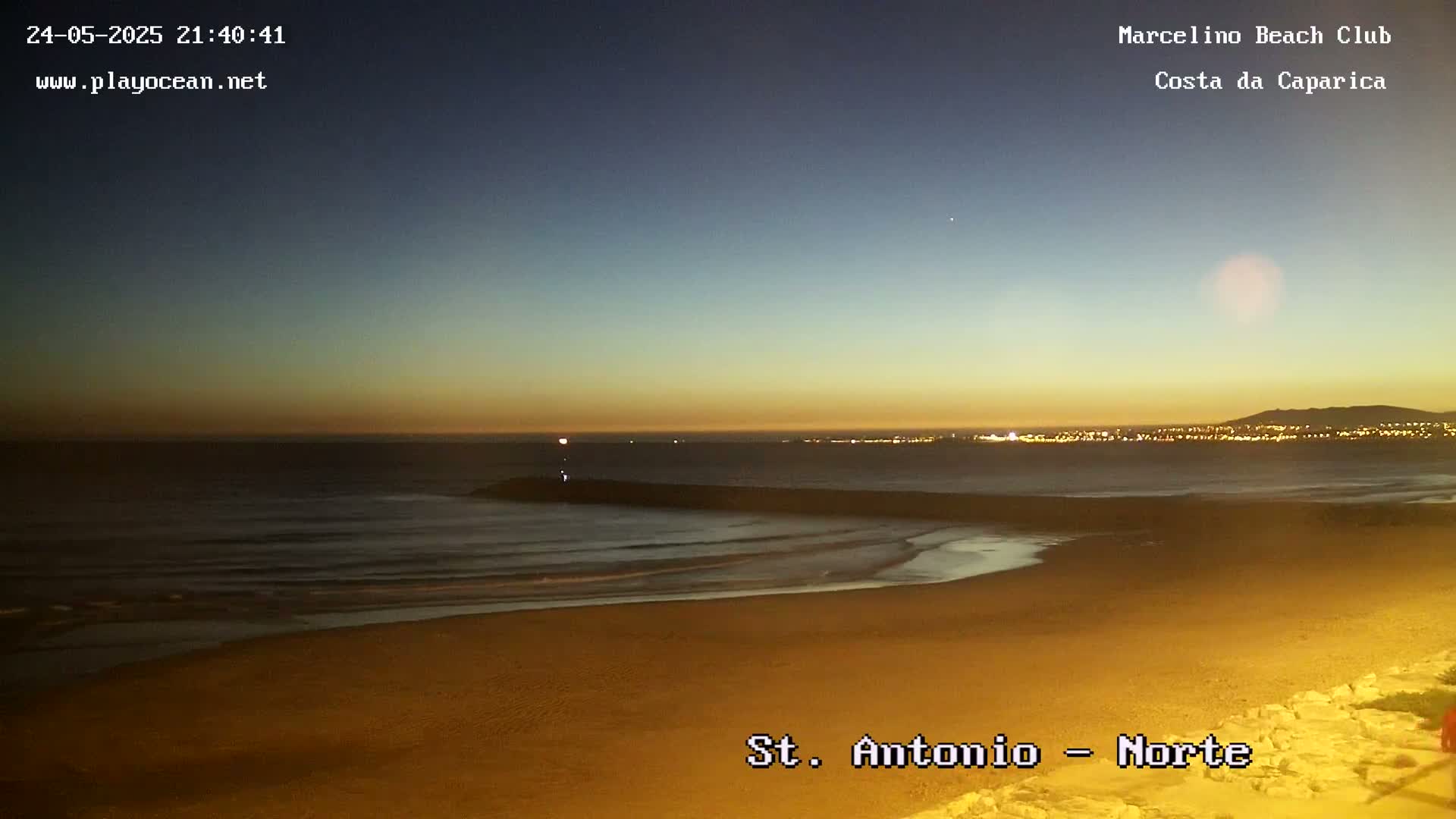 A sandy beach at twilight, with calm waves lapping the shore and a distant city skyline illuminated at night.