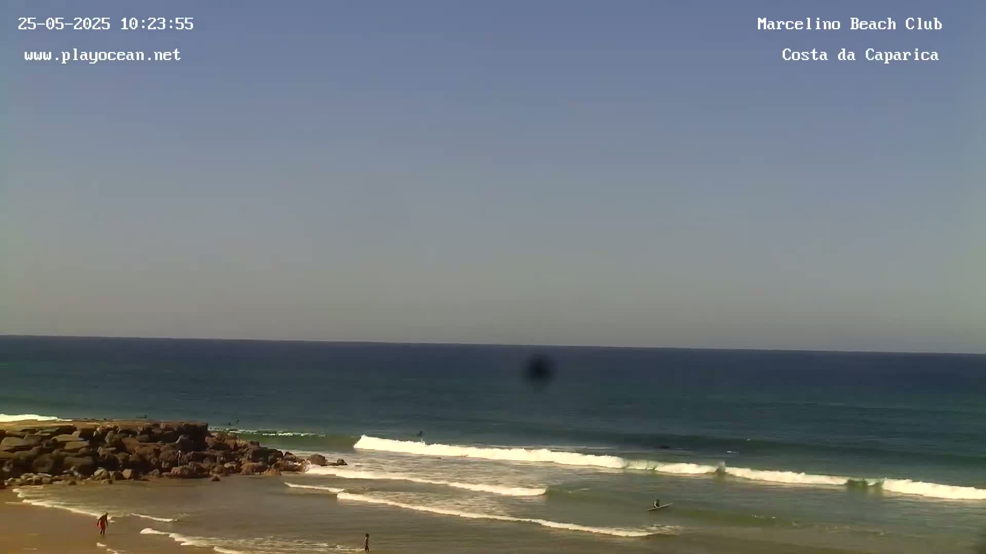 A sandy beach with a rocky outcrop on the left, several people are in the water, some surfing, under a clear blue sky.