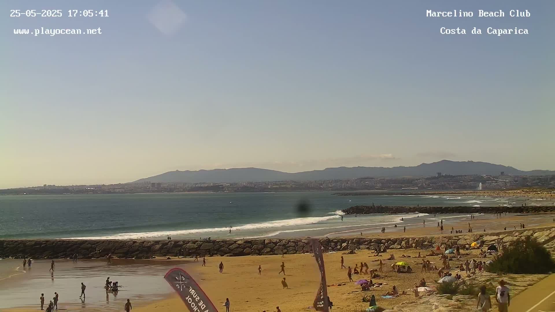 A sunny beach scene shows a calm ocean with small waves, a sandy shore crowded with sunbathers, and a distant city skyline backed by mountains.