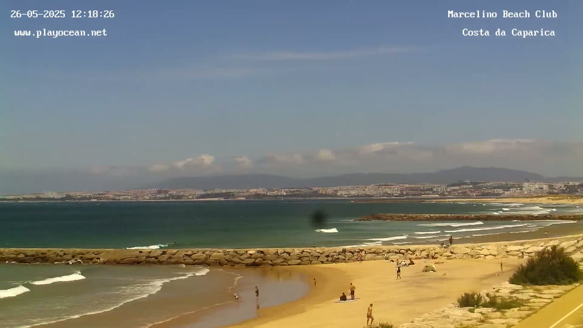 A sandy beach with a few people on it, a calm ocean with some small waves, and a distant city under a partly cloudy sky.