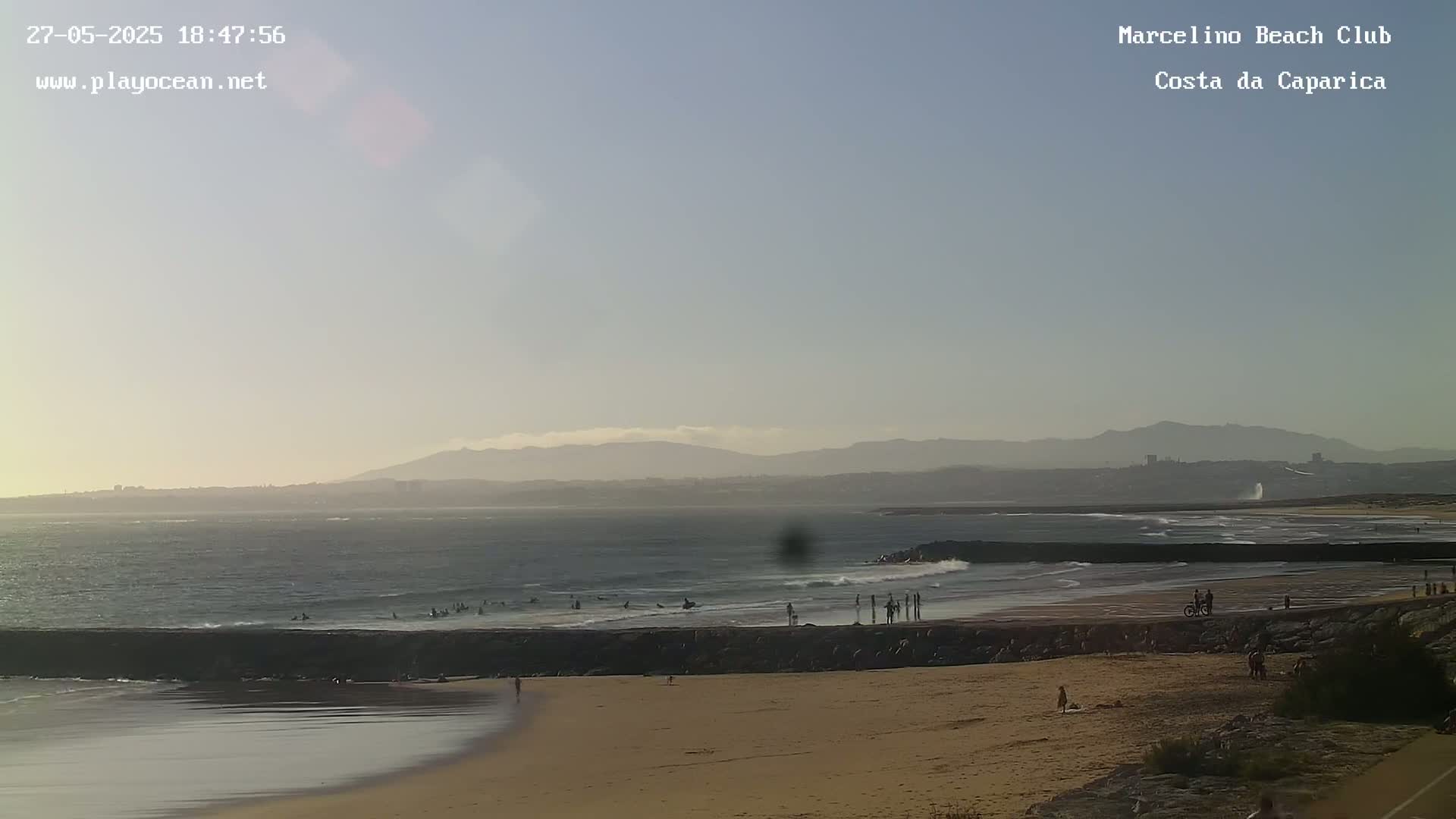 A hazy sunset illuminates a sandy beach with people enjoying the ocean, small waves breaking near a rock jetty, and distant mountains under a clear sky.