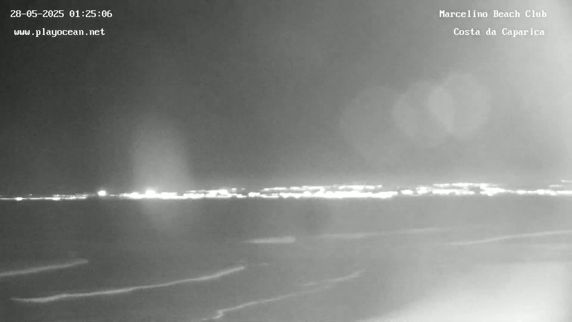 A nighttime, grayscale view of a dark ocean with a distant, faintly lit shoreline under a cloudy sky.