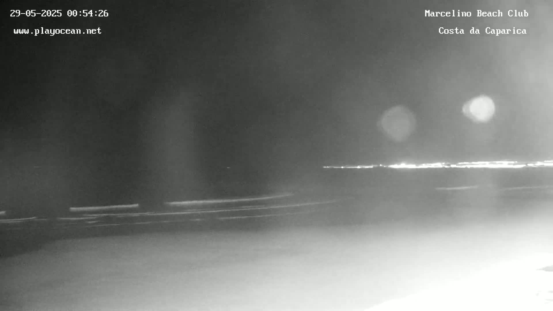 A nighttime, low-light view shows a dark beach with a faint, distant line of lights on the horizon.