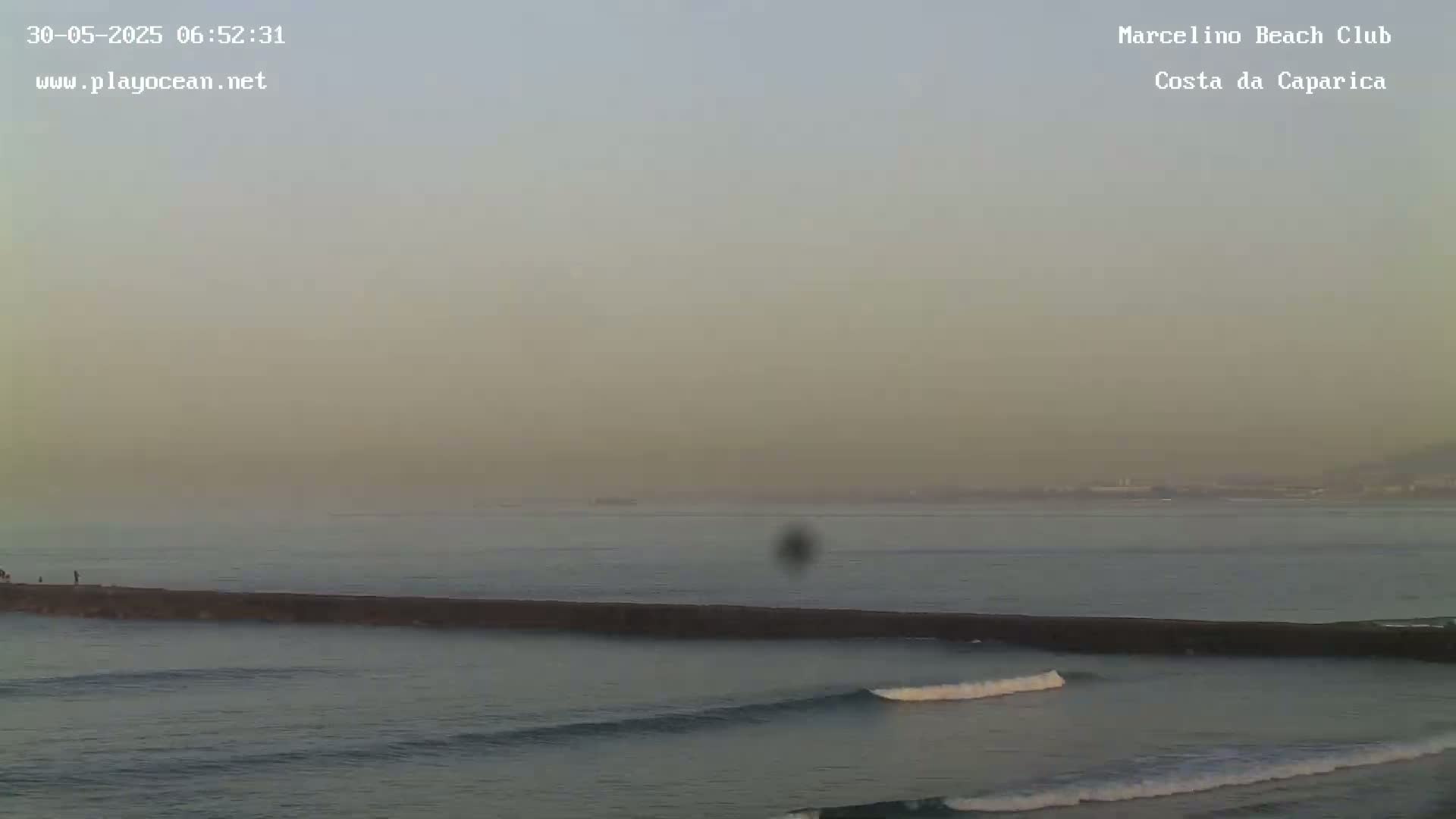 A hazy sunrise reveals a long, dark jetty extending into a calm ocean with small waves breaking near the shore.