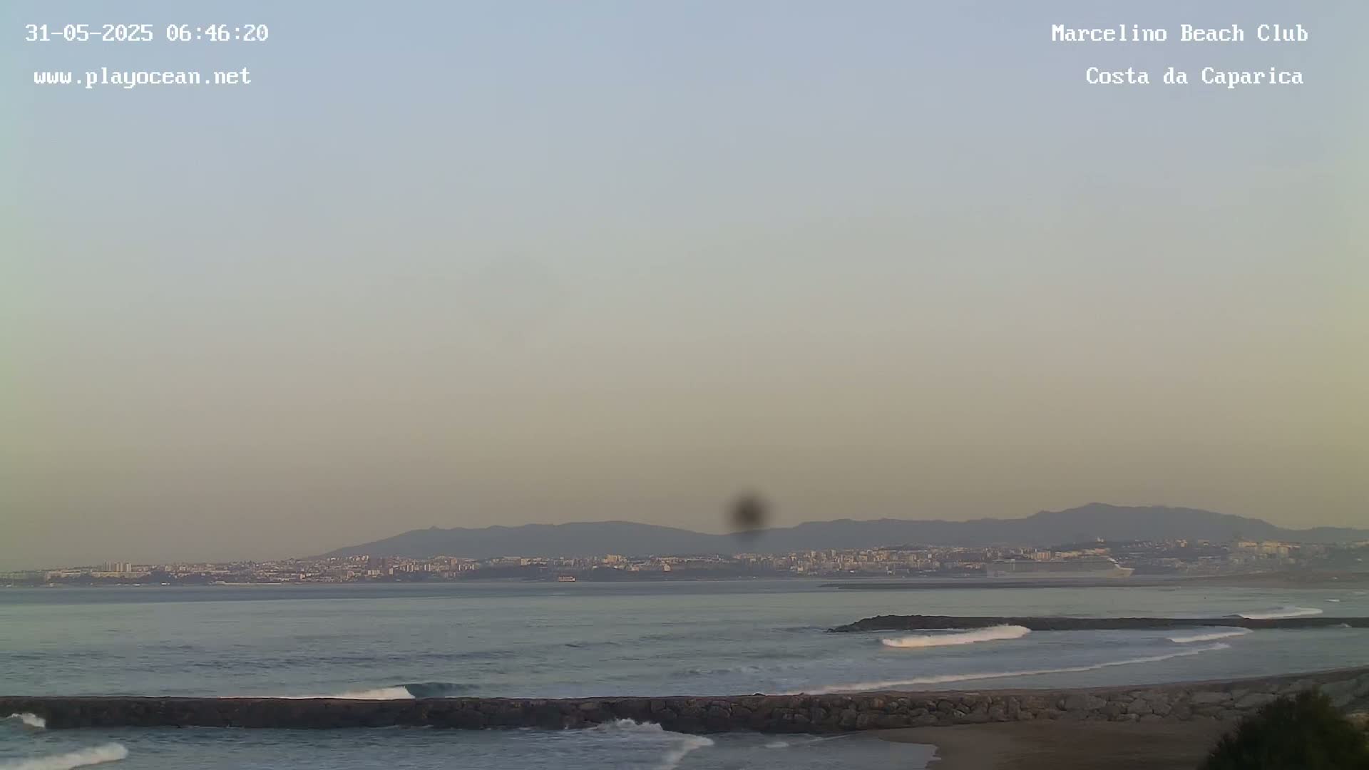 A hazy sunrise reveals a calm ocean with small waves breaking against a rock wall, a distant city skyline, and mountains on the horizon.