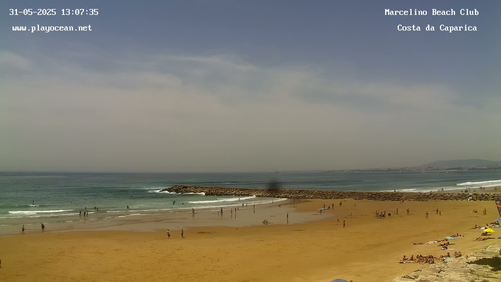 A sandy beach with many people swimming and sunbathing, under a partly cloudy sky, with gentle waves breaking on a rock jetty.