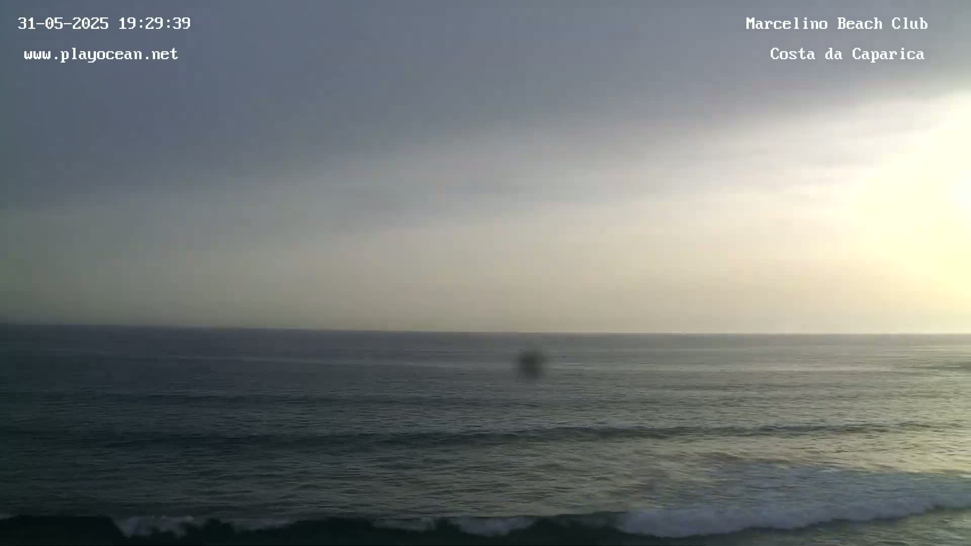 A mostly cloudy, hazy sky overlooks a dark ocean with small waves rolling towards the shore at sunset.