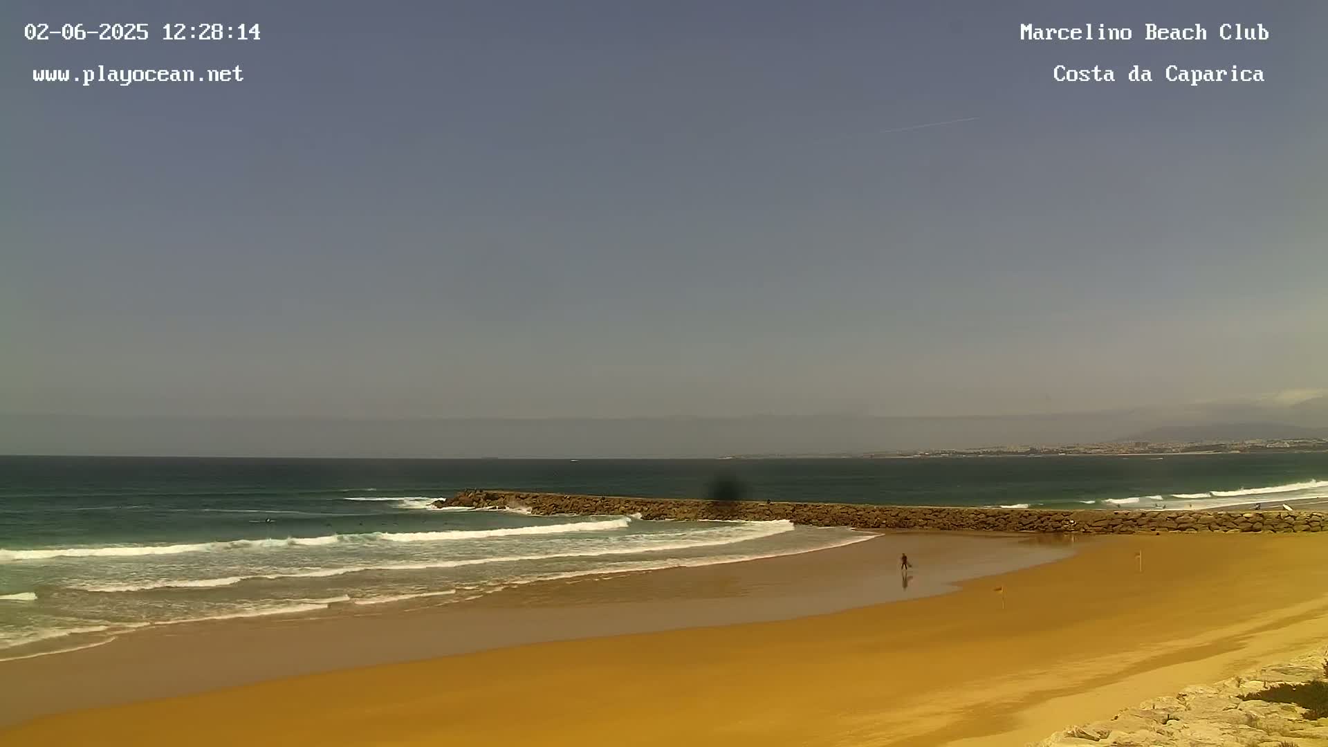A sandy beach with a wave break and several surfers in the ocean under a clear, light-blue sky.