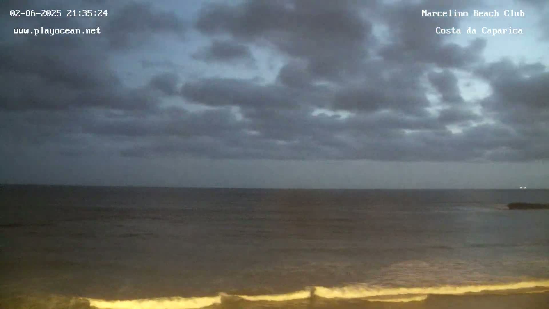 A dark, cloudy evening sky overlooks a calm, dark ocean with gently lapping waves on a beach.
