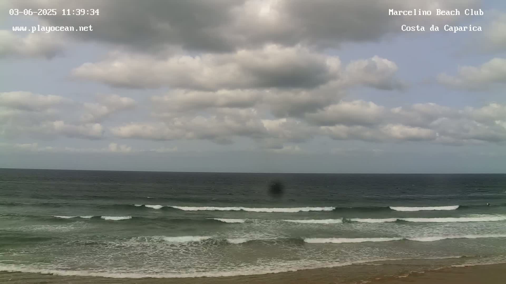 A mostly cloudy sky overlooks a dark-colored ocean with small waves lapping a sandy beach.