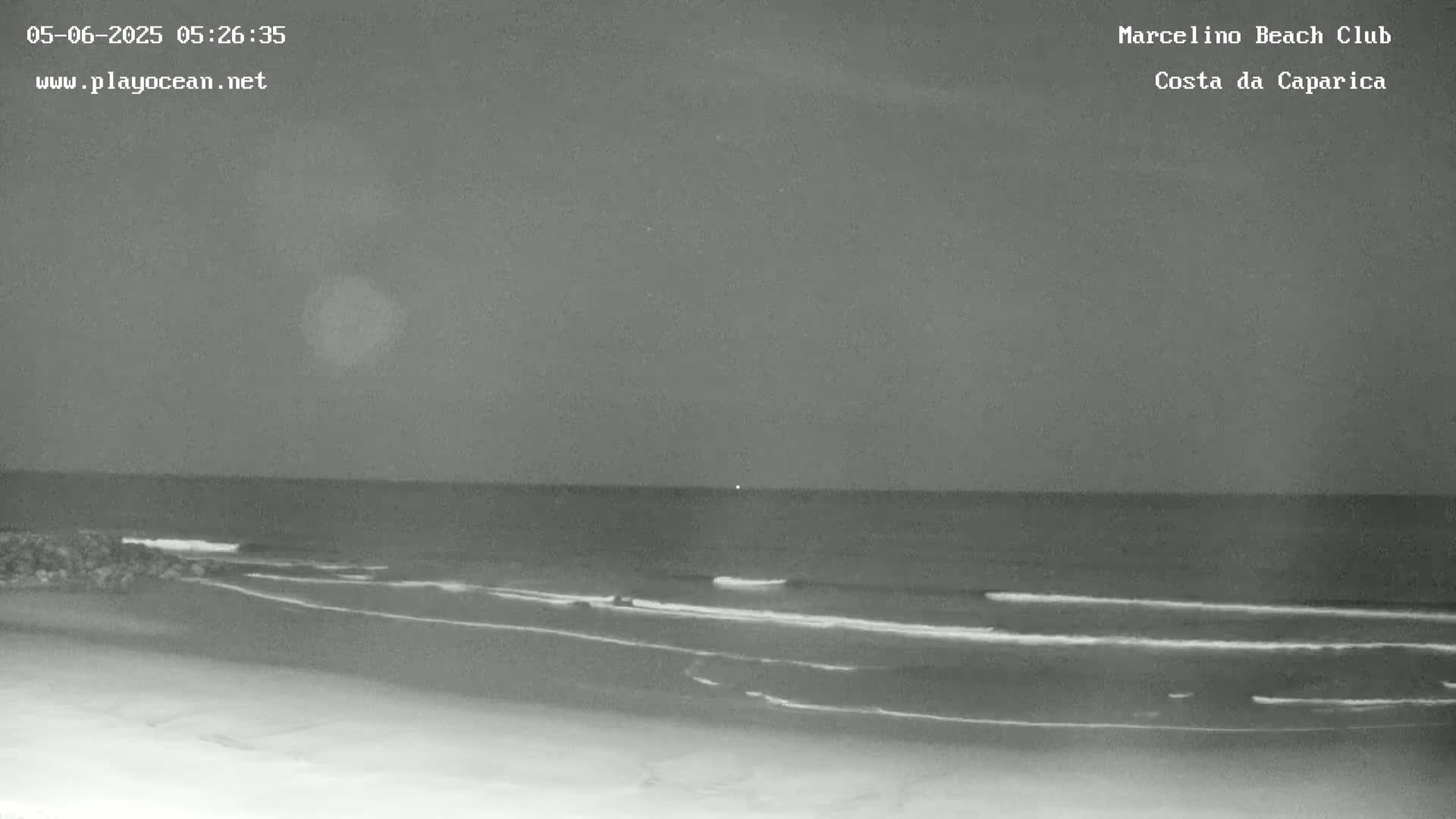 A nighttime, grayscale image shows a dark beach with gentle waves lapping the shore under a cloudy sky.