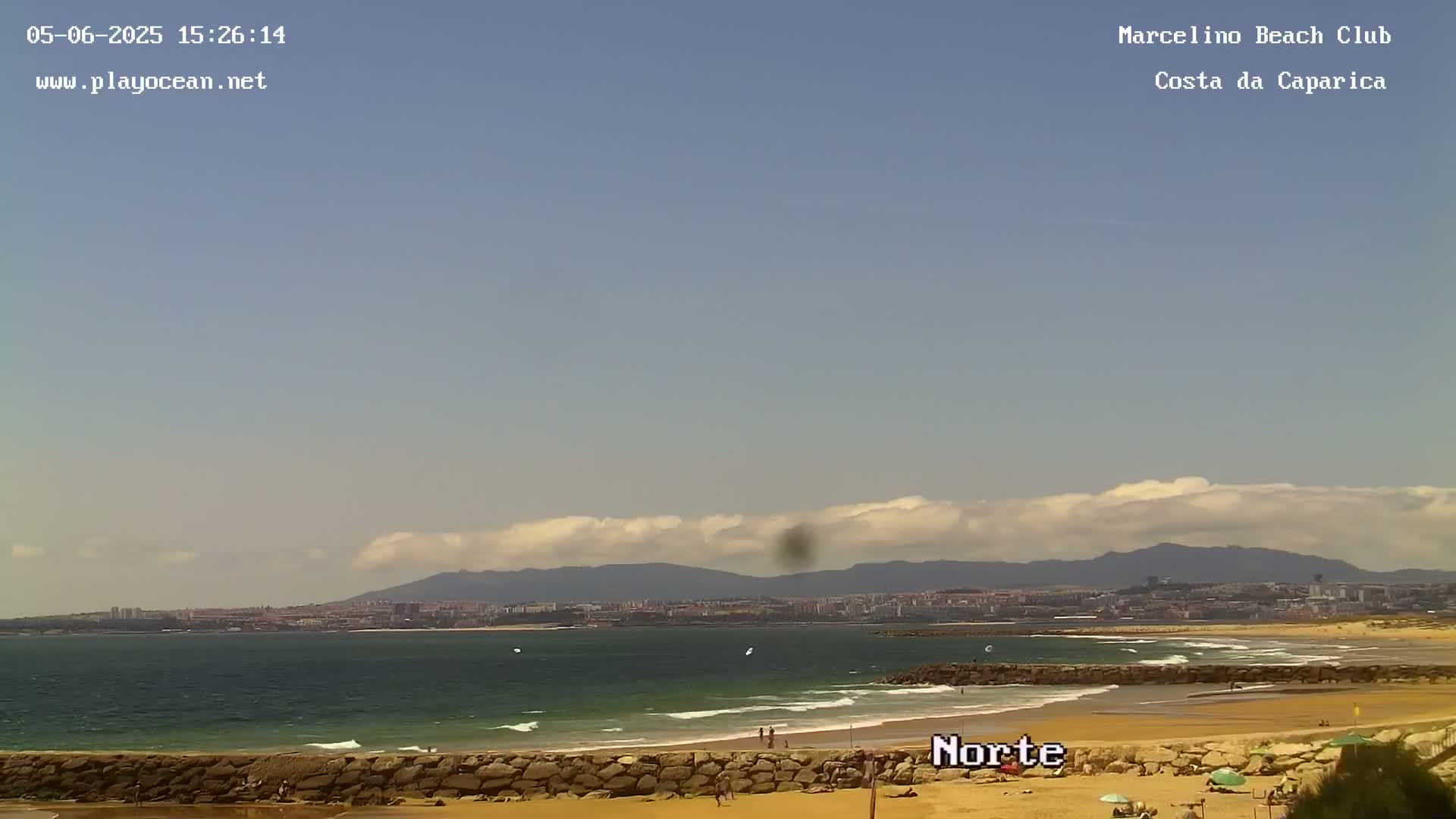 A mostly sunny day at a sandy beach with calm ocean waves, a distant city skyline, and rolling hills under a partly cloudy sky.