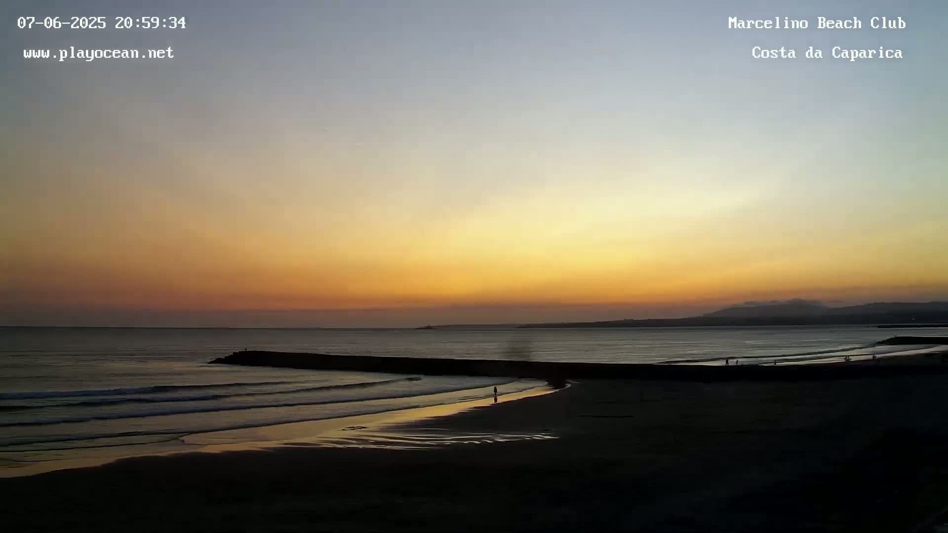 A beach at sunset shows a calm ocean with small waves lapping the shore under a yellow and orange sky.