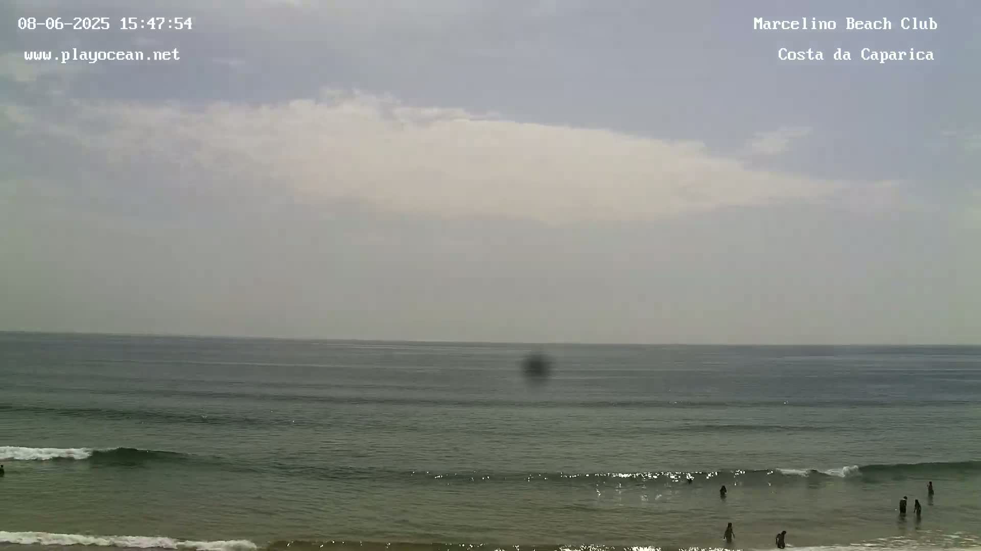 A mostly cloudy day at a beach shows a calm ocean with several people in the water near the shore.
