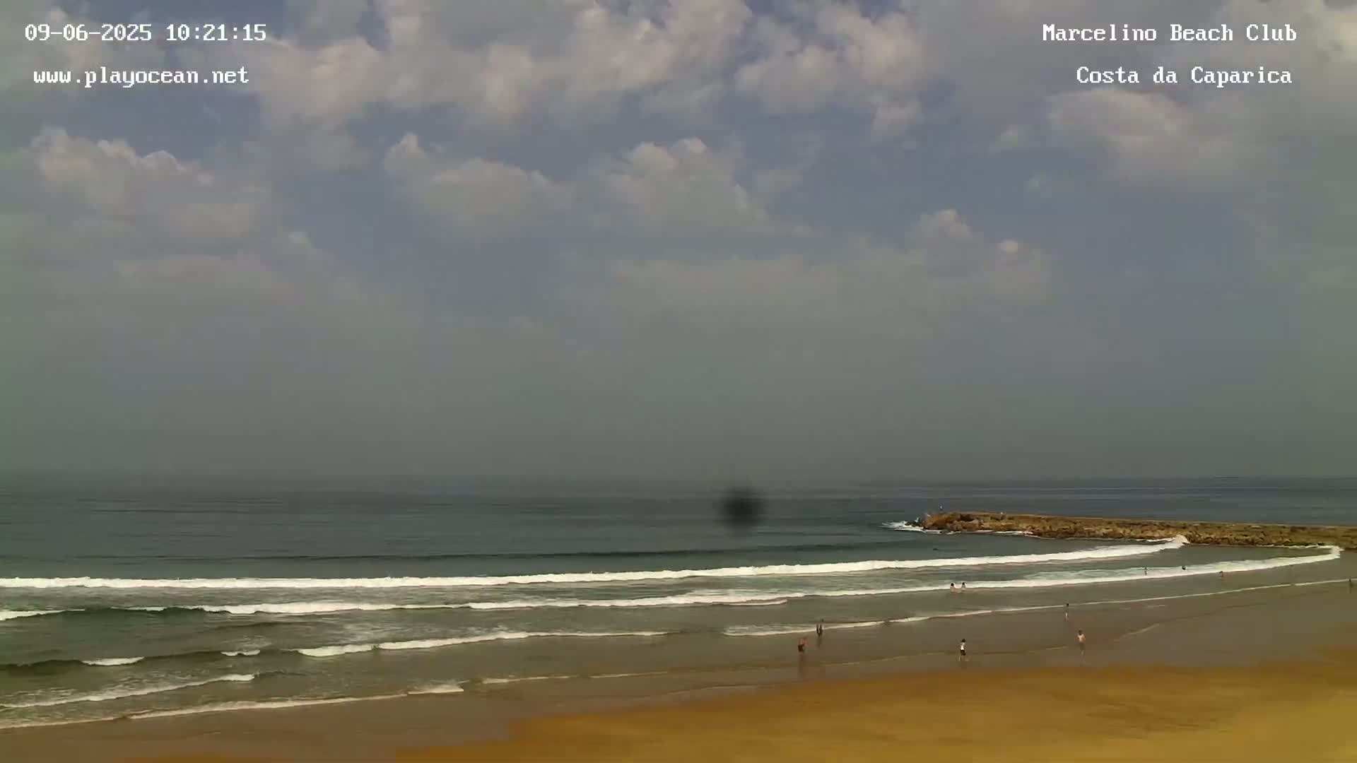 A hazy, partly cloudy day at a beach with small waves breaking near a rocky jetty, and a few people are visible on the sand.
