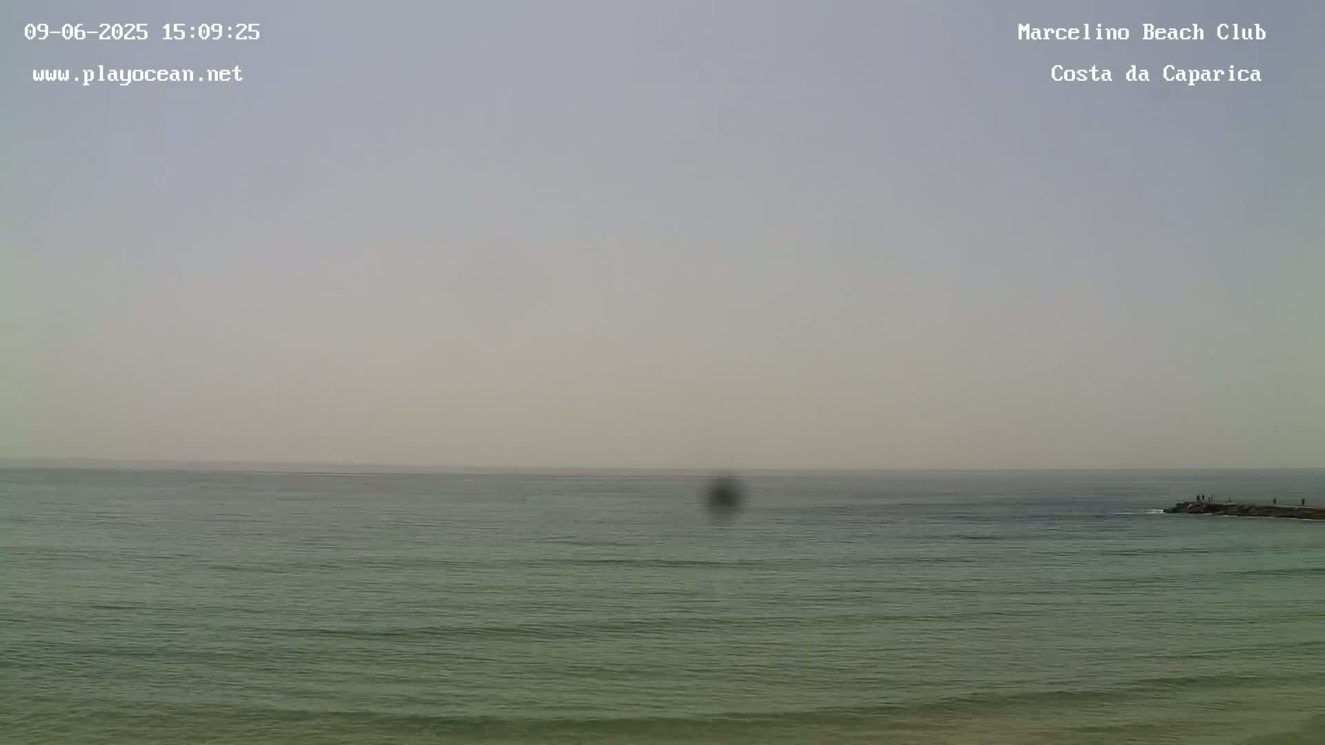 A hazy, calm ocean stretches to a pale horizon, with a few people visible on a distant rock jetty.